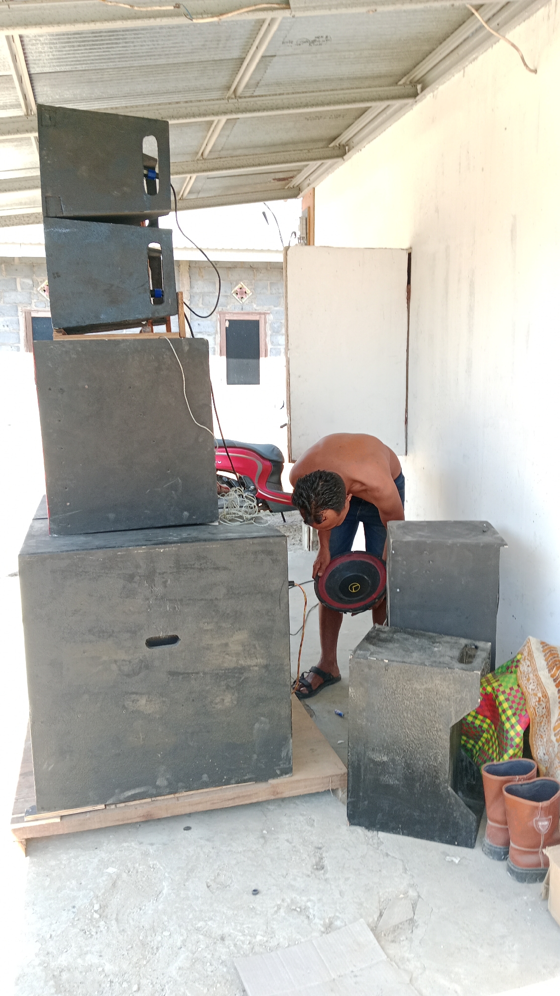 In Karang Indah, Indonesia, a shirtless man is seen in a partially covered outdoor area, possibly a workshop or storage space. He is bending over, holding a speaker component with a red trim, and appears to be in the process of assembling or repairing audio equipment. Several large, black speaker cabinets are stacked and arranged around him. A red scooter is partially visible behind the speaker stacks, and coiled wires are scattered on the ground. The environment appears to be utilitarian, with a corrugated metal roof and cinder block walls visible in the background, suggesting a practical, possibly residential or small business setting. Two brown boots and a colorful fabric are placed near the speaker cabinets on the right side of the image. The ground is concrete, with some debris present.