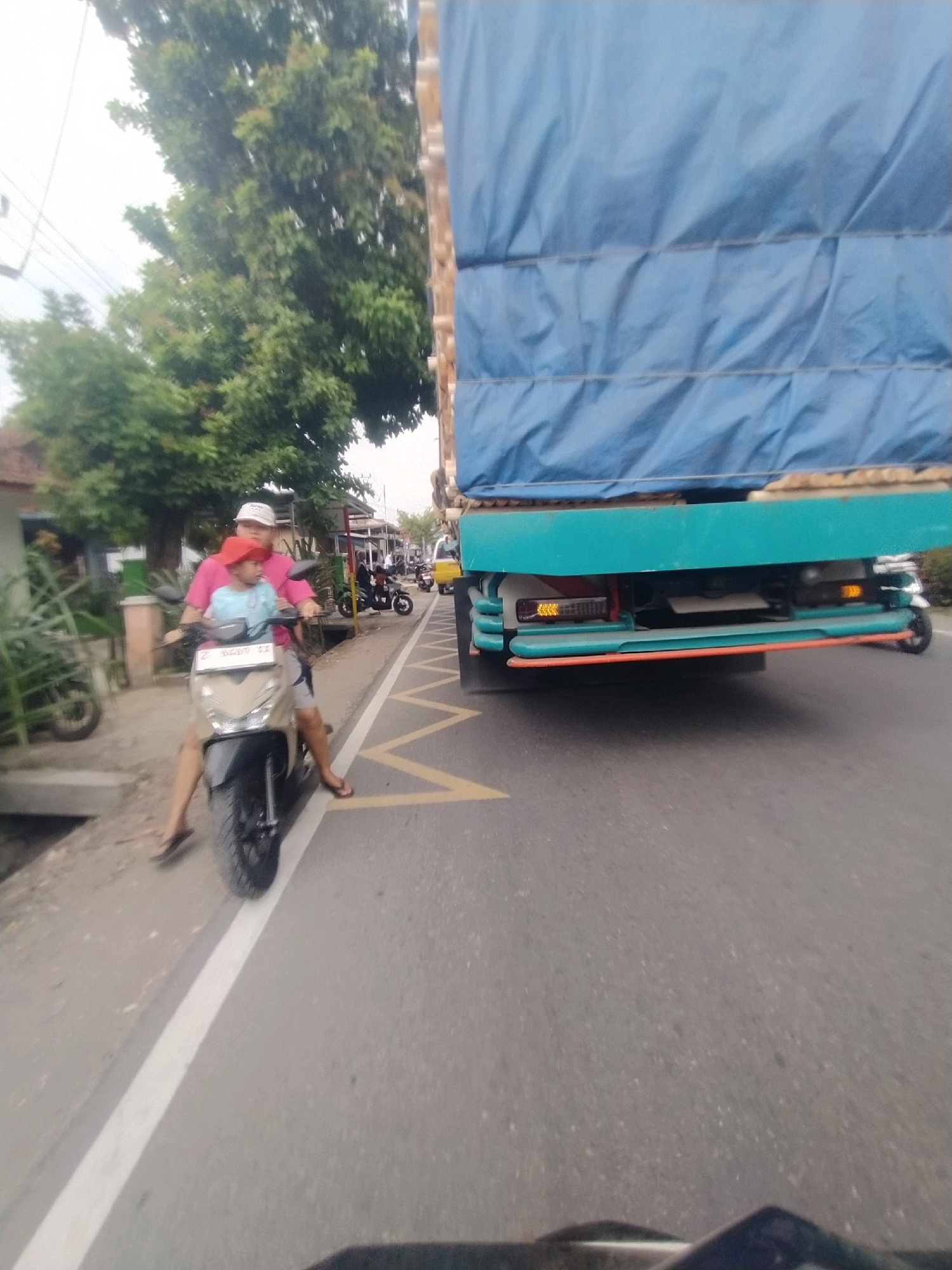 In Desa Karangbenda, Indonesia, a person is riding a scooter on a paved road. The rider, wearing a pink shirt and a white cap, is carrying a child in a light blue shirt and a red hat on the front of the scooter. The scooter is a light brown color and is positioned on the left side of the road, with a white line marking the lane. In the background, there are trees and some buildings. To the right of the scooter, a large blue-covered truck is visible, parked or moving on the road. The truck has a teal-colored frame and orange accents. The sky appears overcast.