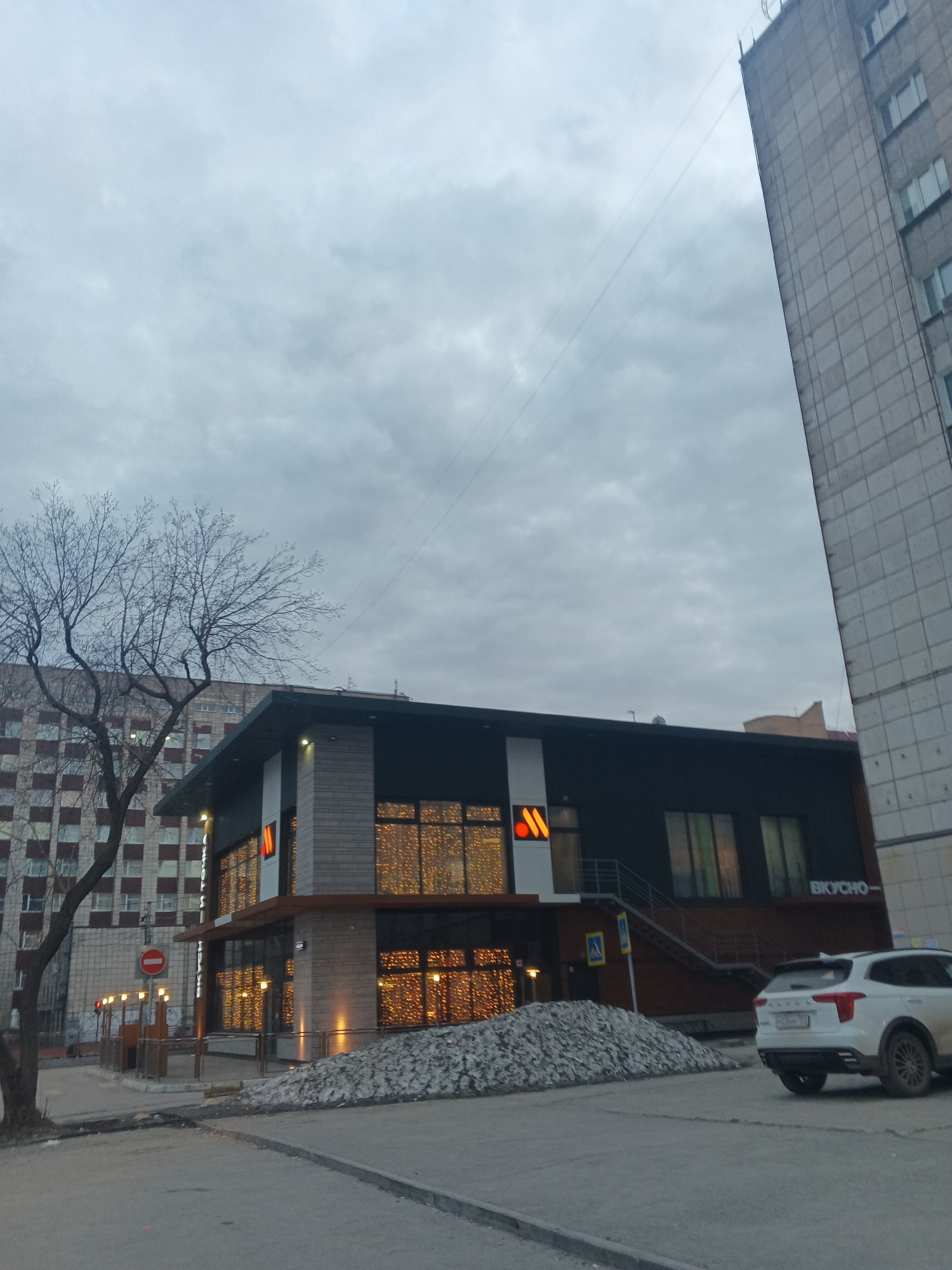 The image captures a street scene in Perm, Russia, featuring a "Вкусно – и точка" restaurant (formerly McDonald's). The building is two-storied, with an orange "M" logo visible on the facade. The windows are adorned with warm-toned lights, contrasting with the grey overcast sky. A white SUV is parked nearby. In the foreground, a bare tree stands to the left, and a large pile of debris or construction material lies in front of the restaurant. A tall apartment building borders the right side of the frame. Street signs and other urban infrastructure are also present. The lighting suggests it is either early morning or late evening.