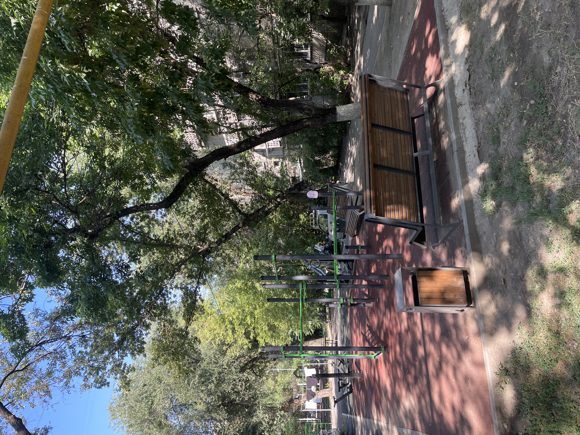 This is an outdoor image of a street workout area in Almaty, Kazakhstan, located in a residential courtyard. The ground is covered with a red rubber mat, and there are various exercise equipment structures made of metal pipes with green accents, including pull-up bars and parallel bars. In the foreground, there are two wooden benches with metal frames, one facing the workout equipment and the other facing away. A wooden and metal trash can is situated next to the bench in the foreground. Several mature trees with lush green foliage cast dappled shadows on the ground. In the background, a multi-story apartment building with visible windows and air conditioning units is visible, partially obscured by trees. There is a white car parked on the right side of the image. The sky is clear and blue. The image appears to have been taken during the daytime.