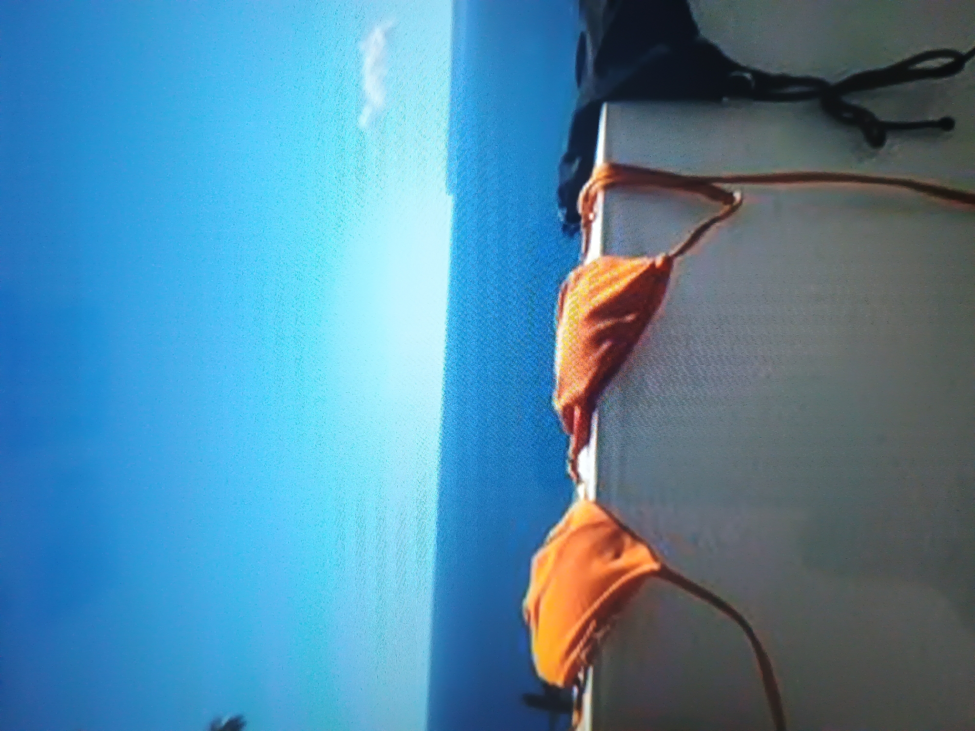 A high-angle outdoor photograph captures a broad expanse of deep blue ocean meeting a lighter blue sky at the horizon line. A single, small white cloud is visible in the upper right quadrant of the sky. In the foreground, a light-colored, horizontal wall or railing is prominently featured. Draped over this wall are multiple items of swimwear. On the left, an orange bikini top is visible, with its straps extending downwards along the wall. Adjacent to it, another orange garment, likely a matching bikini top or a component of swimwear, is also draped. To the far right, a dark, possibly black, piece of clothing, consistent with swimwear, hangs over the wall. No individuals are present in the scene. The lighting indicates a bright, sunny day. The location is Unknown, Unknown.