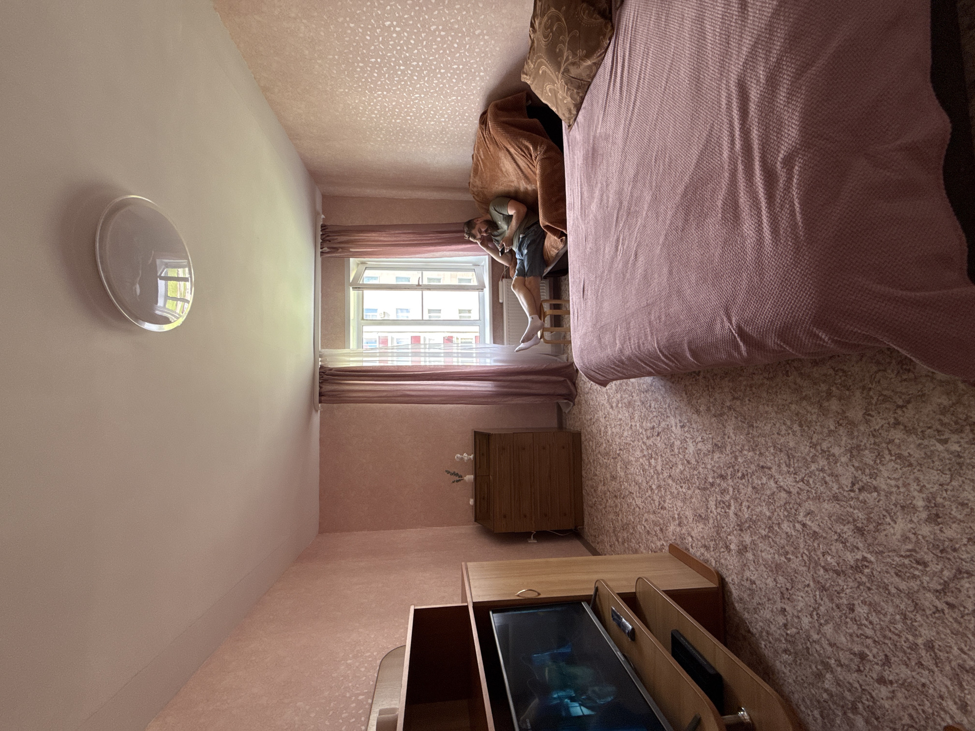 The image shows a bedroom in an apartment in Valday, Russia. A man is sitting on a couch, looking at his phone. The room has pink walls and a patterned carpet. There is a bed, a dresser, and a television. The window is open, and curtains are drawn.