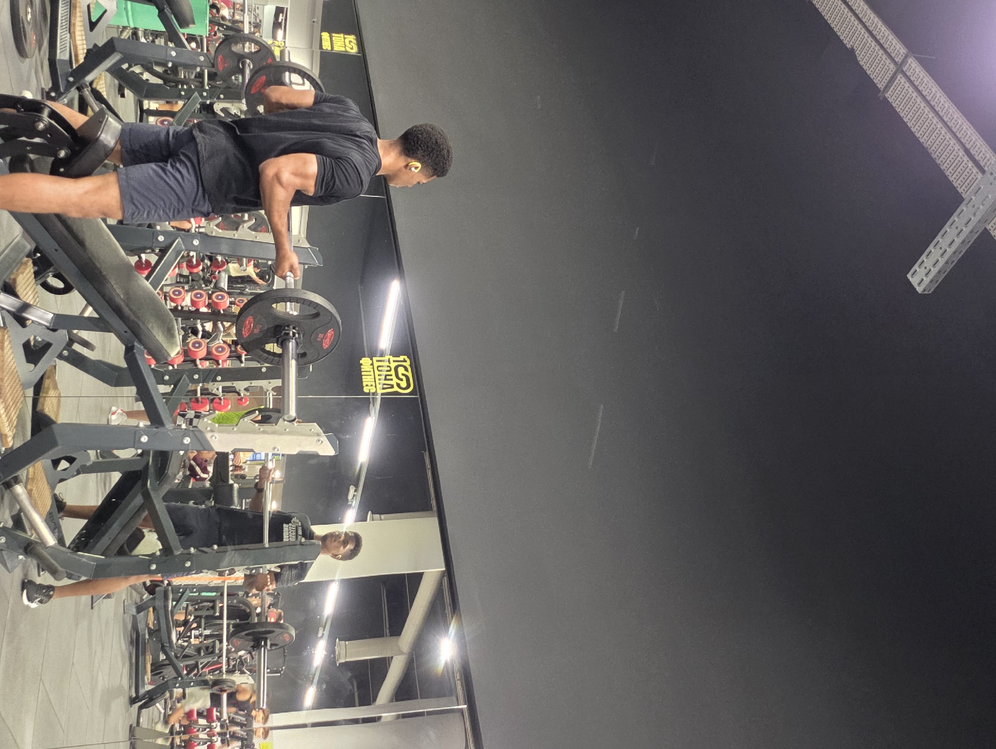 The image depicts the interior of a gym, likely located in Varna, Bulgaria, evidenced by the "100 HA fitness" logo, a prominent fitness chain in Varna. Two young men are engaged in weightlifting. In the foreground, a man with dark skin and short, curly hair is performing bicep curls with a barbell. He is wearing a black t-shirt and dark shorts. In the background, another man, with lighter skin and short hair, is standing with a barbell. He is wearing a black t-shirt and dark shorts. The gym environment is characterized by various weightlifting equipment, including weight benches and racks, with mirrors reflecting the scene. The lighting appears to be artificial, common in indoor fitness facilities.