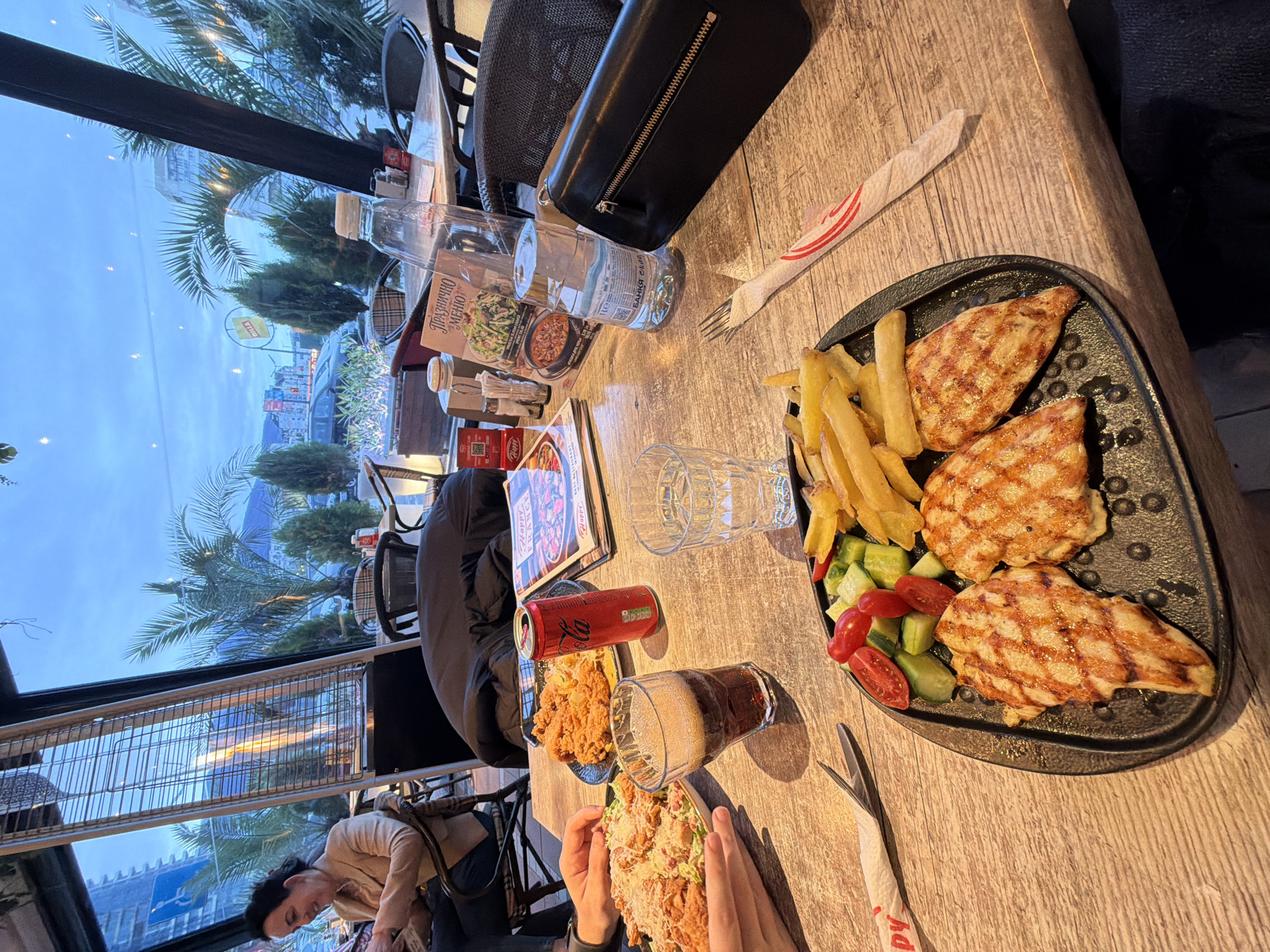 This photograph captures a dining experience at a restaurant, likely during the evening or dusk, given the twilight blue sky visible through the large window. The image is taken from a perspective looking down at a wooden table laden with food and drinks.
In the foreground, a person's hands are visible, holding a large plate or bowl containing what appears to be a generous serving of a dish with fried chicken or a large burger/sandwich, topped with a creamy sauce and possibly coleslaw. To the right, on a black platter, are three pieces of grilled chicken, alongside a portion of golden French fries and a fresh side salad composed of diced cucumbers and cherry tomatoes. A white napkin with a red design (partially showing "Happy") and a fork are placed next to this plate.
Various drinks are spread across the table, including two clear plastic bottles of water (one clearly marked "БАНКЯ" (Bankya), a Bulgarian mineral water brand), a red Coca-Cola can, and a glass filled with a dark, carbonated beverage, possibly cola or beer. A clear, empty glass is also present.
Other objects on the table include a black leather purse, a menu with "ПРАЗНИЧНО МЕНЮ" (Praznichno Menyu - Festive Menu) visible in Cyrillic, and small condiment shakers.
Through the expansive window on the left side of the frame, an outdoor scene is visible. This includes palm trees and other greenery, urban buildings, and what appears to be an outdoor heater, suggesting an open-air or semi-open dining area. A yellow sign is partially visible in the distance. Another person is seated at the table on the far left, leaning forward, wearing a light-colored jacket. The overall atmosphere suggests a casual and enjoyable meal. The visible Bulgarian text on the water bottle and menu confirms the location context of Sofia, Bulgaria.