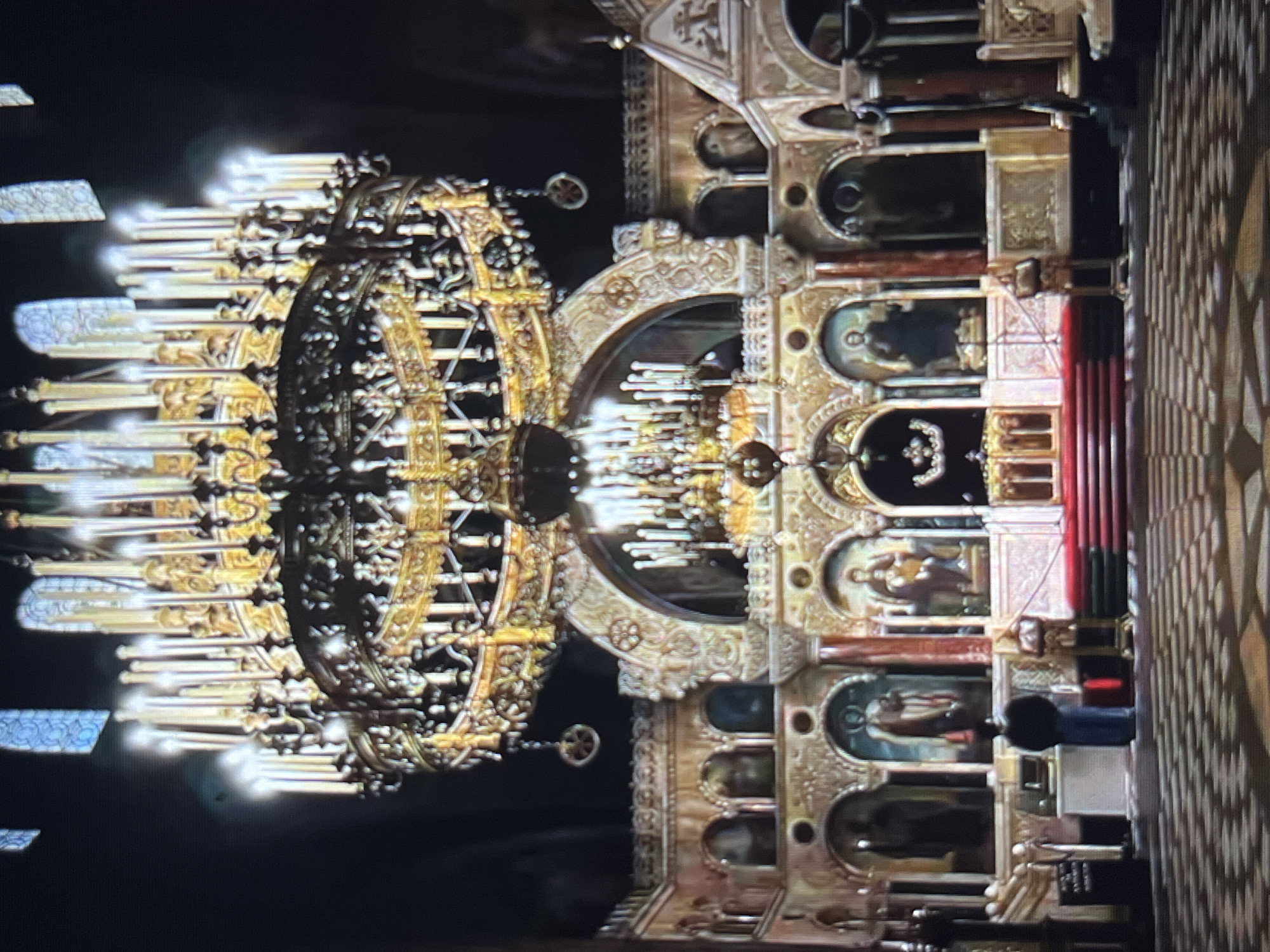 The image shows the interior of a church or cathedral in Tinaquillo, Venezuela, featuring ornate gold detailing. A large, multi-tiered chandelier with numerous lights dominates the left side of the frame. The right side displays a gold-colored wall with multiple niches containing religious icons or artwork. The floor shows a patterned design. A person stands near the right side, in front of the gold wall.