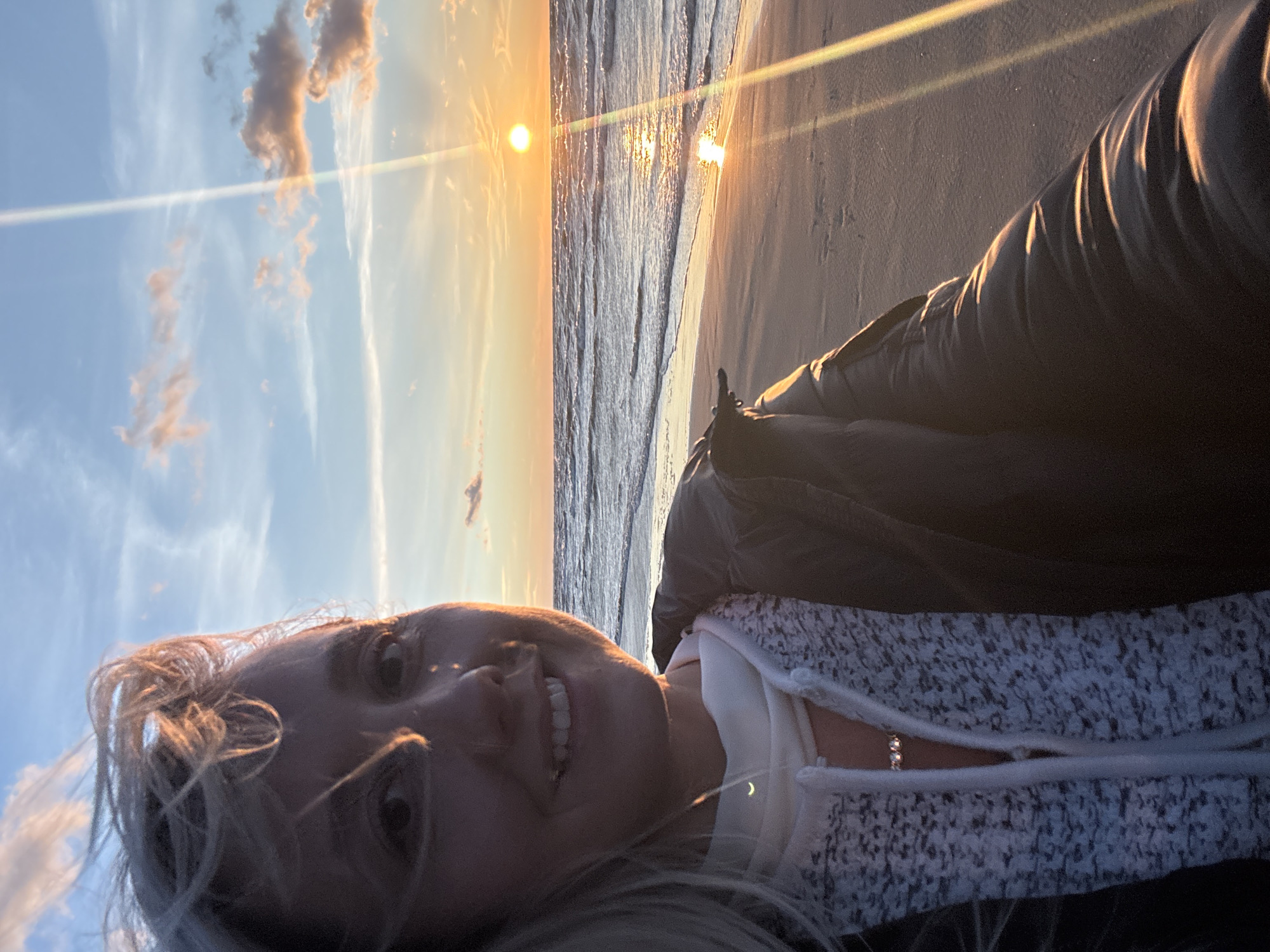 A woman is taking a selfie on a beach at sunset. The woman has blonde hair, and is smiling at the camera. She is wearing a black jacket over a white hoodie. The sun is setting over the water, casting a warm glow on the scene. The waves are gently crashing on the shore. The image appears to have been taken in Jūrmala, Latvia, as evidenced by the distinct characteristics of the Baltic Sea coast and the sky coloration typical of the region during sunset.
