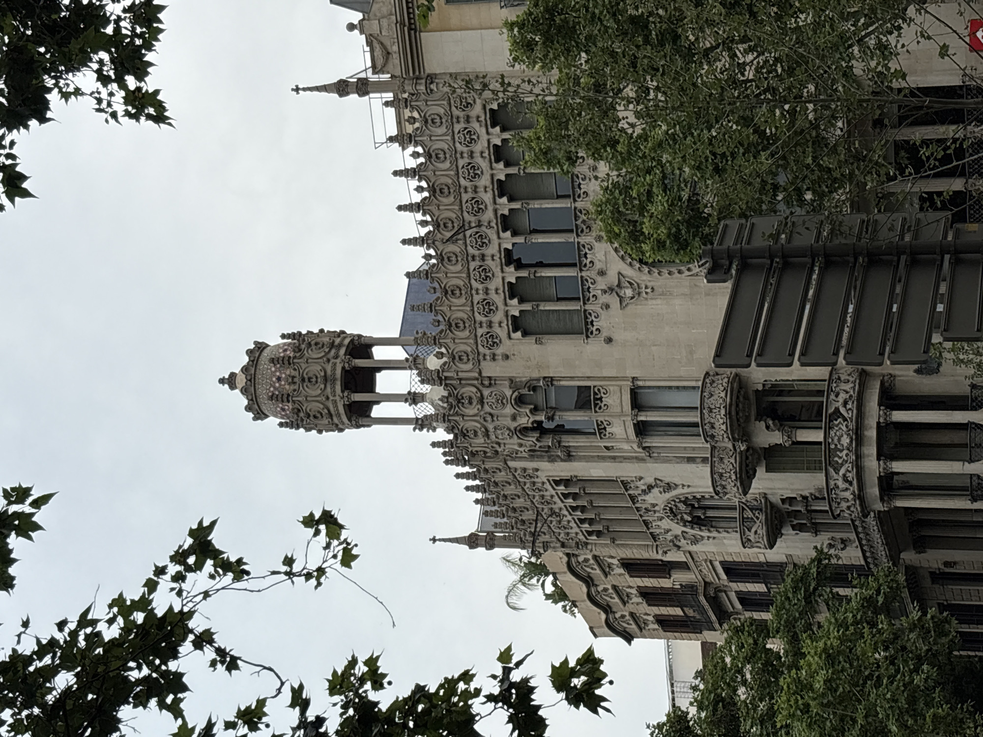 The image shows a detailed view of Casa Comalat, a modernist building located in Barcelona, Spain, designed by Salvador Valeri i Pupurull. The building showcases intricate architectural details, including ornamented balconies, windows, and a distinctive roof with a turret. The facade is primarily off-white, contrasting with the darker tones of the windows and the lush greenery of trees in the foreground and background. The scene is captured on a slightly overcast day, which diffuses the light and accentuates the building's sculptural elements.