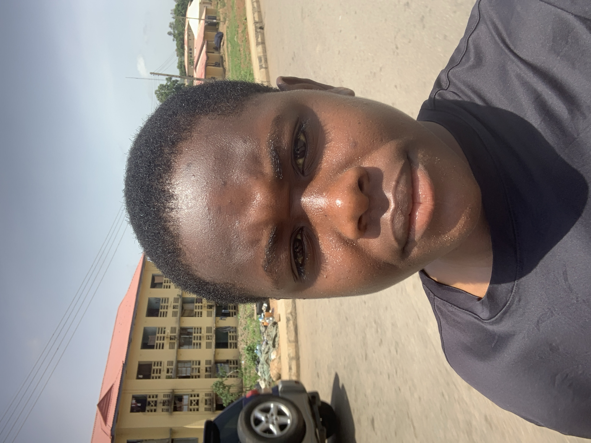 A close-up selfie taken by a young individual outdoors in Jos, Nigeria. The individual, with dark skin and short dark hair, is wearing a dark t-shirt. Their face, glistening with sweat, is prominently featured, with their eyes looking directly at the camera. In the background, a yellow multi-story building with a red roof is visible, along with other smaller buildings and vegetation. A dark-colored SUV is parked to the left of the frame, and the ground appears to be a paved road. The sky is light blue with a few scattered clouds.