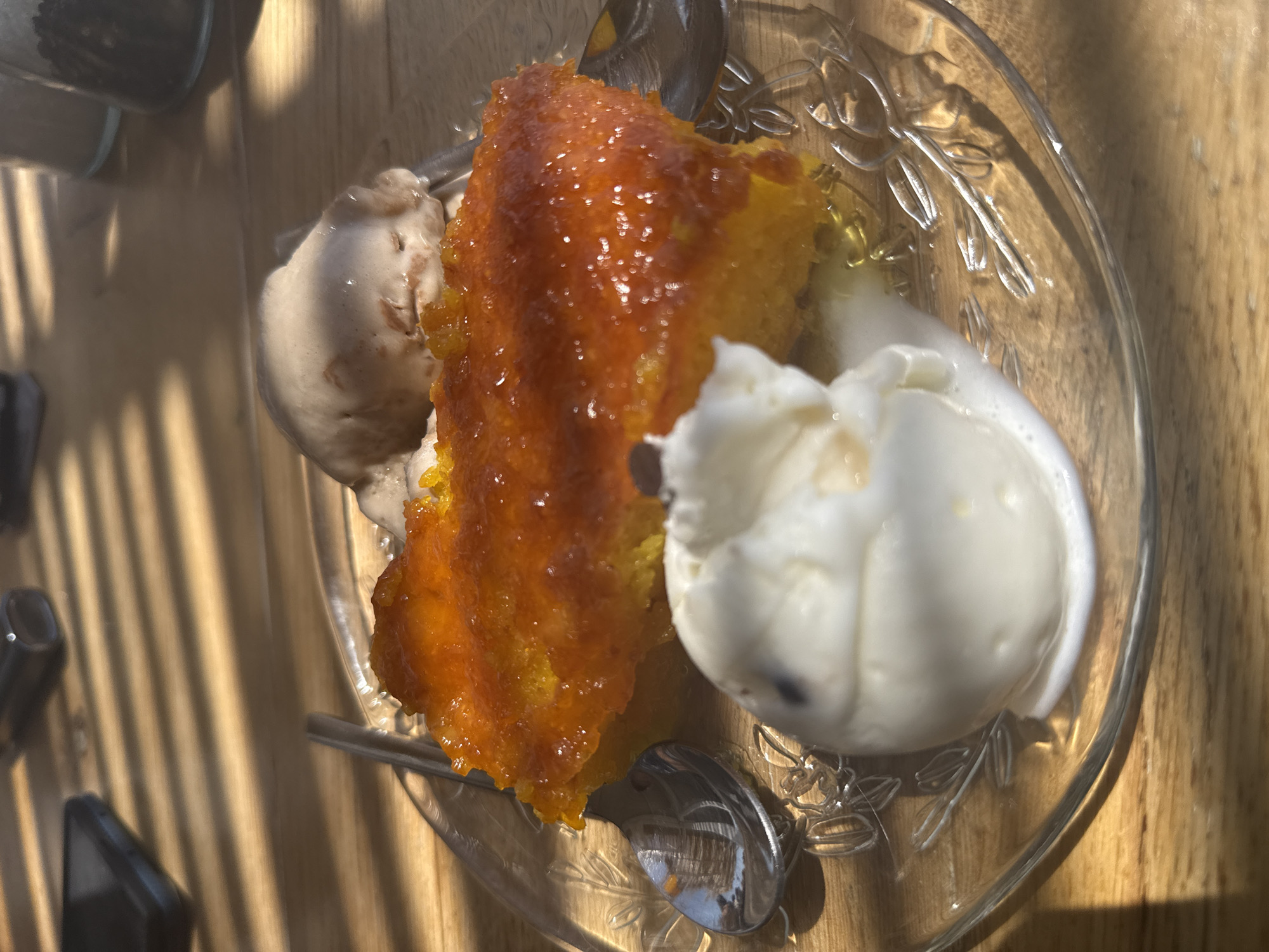 A close-up, high-angle shot shows a slice of vibrant orange cake, possibly pumpkin or sweet potato, topped with a glistening, syrupy glaze. Two scoops of ice cream, one appearing to be vanilla and the other a lighter brown flavor, melt slightly next to the cake on a clear glass plate with a floral design. The plate rests on a light-colored wooden surface, possibly a table, which is illuminated by dappled sunlight casting strong shadows from diagonal blinds or slats. In the background, out of focus, are what appear to be two glass jars. The scene suggests a dessert being served, likely in a cafe or restaurant setting in Λάκωνες, Greece, given the provided context.