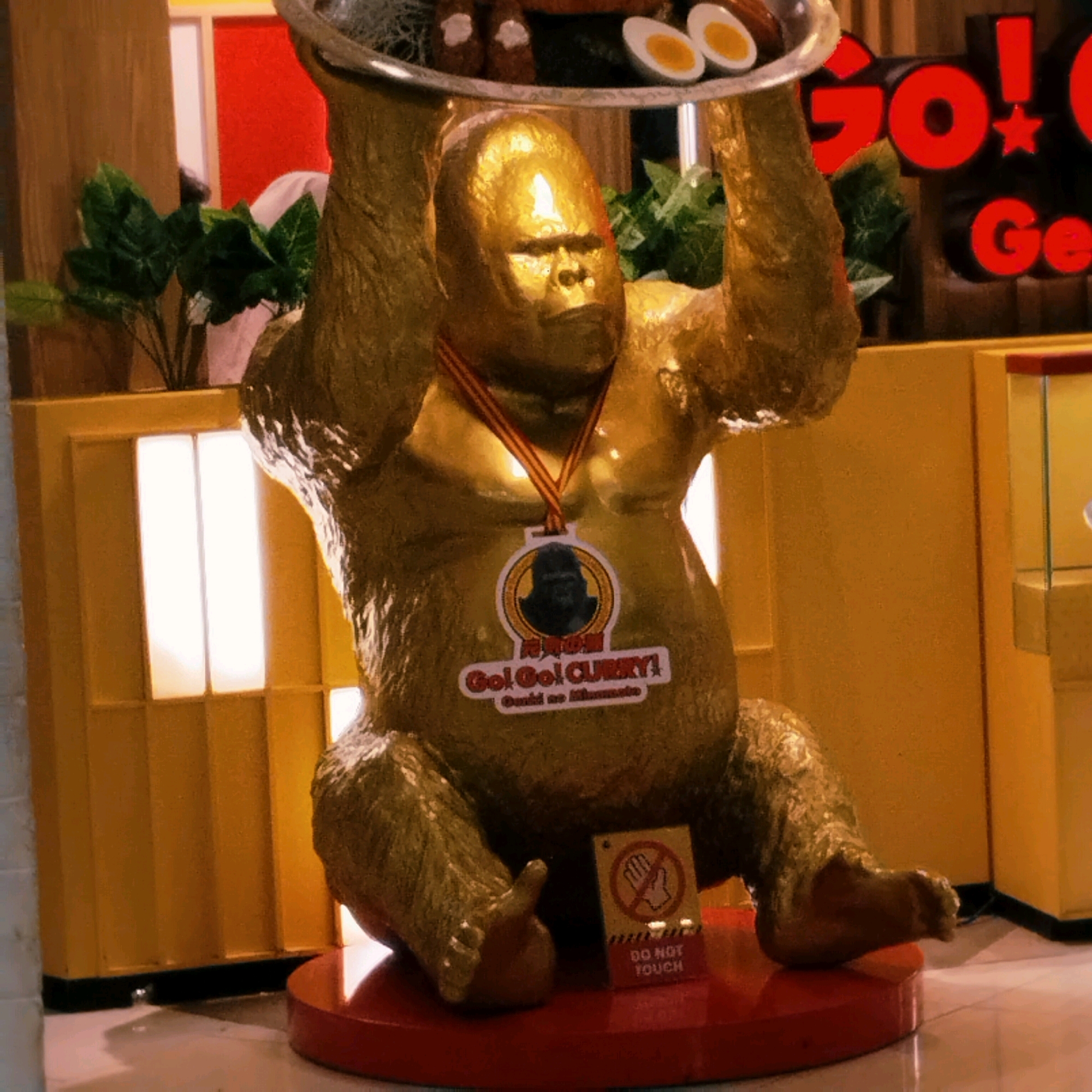 A life-size, golden statue of a gorilla is the primary subject of this image, positioned centrally in what appears to be a restaurant or shop in Surabaya, Indonesia. The gorilla is depicted sitting down, holding a large silver platter above its head. The platter contains food items, including what look like hard-boiled eggs and possibly some meat dishes. The statue wears a red and yellow striped ribbon around its neck, and a circular emblem with a gorilla's silhouette and the text "Go! Go! CURRY!" is affixed to its chest. To the lower right of the statue, a small yellow and red sign displays a hand symbol within a circle and the text "DO NOT TOUCH."
The background suggests an indoor commercial setting. To the left of the gorilla, there are tall, yellow vertical panels with illuminated rectangular sections that resemble windows or display lights. Behind the gorilla, a red wall is visible, and on the right side of the image, there is a display case with glass panels, showcasing items within. Further to the right, bold red lettering on a dark background spells out "Go! Go!" and "Ge". Green artificial plants are strategically placed around the statue, adding a touch of nature to the artificial environment. The floor is a light-colored tiled surface. The lighting is somewhat dim, with bright spots on the gorilla's face and the yellow panels.