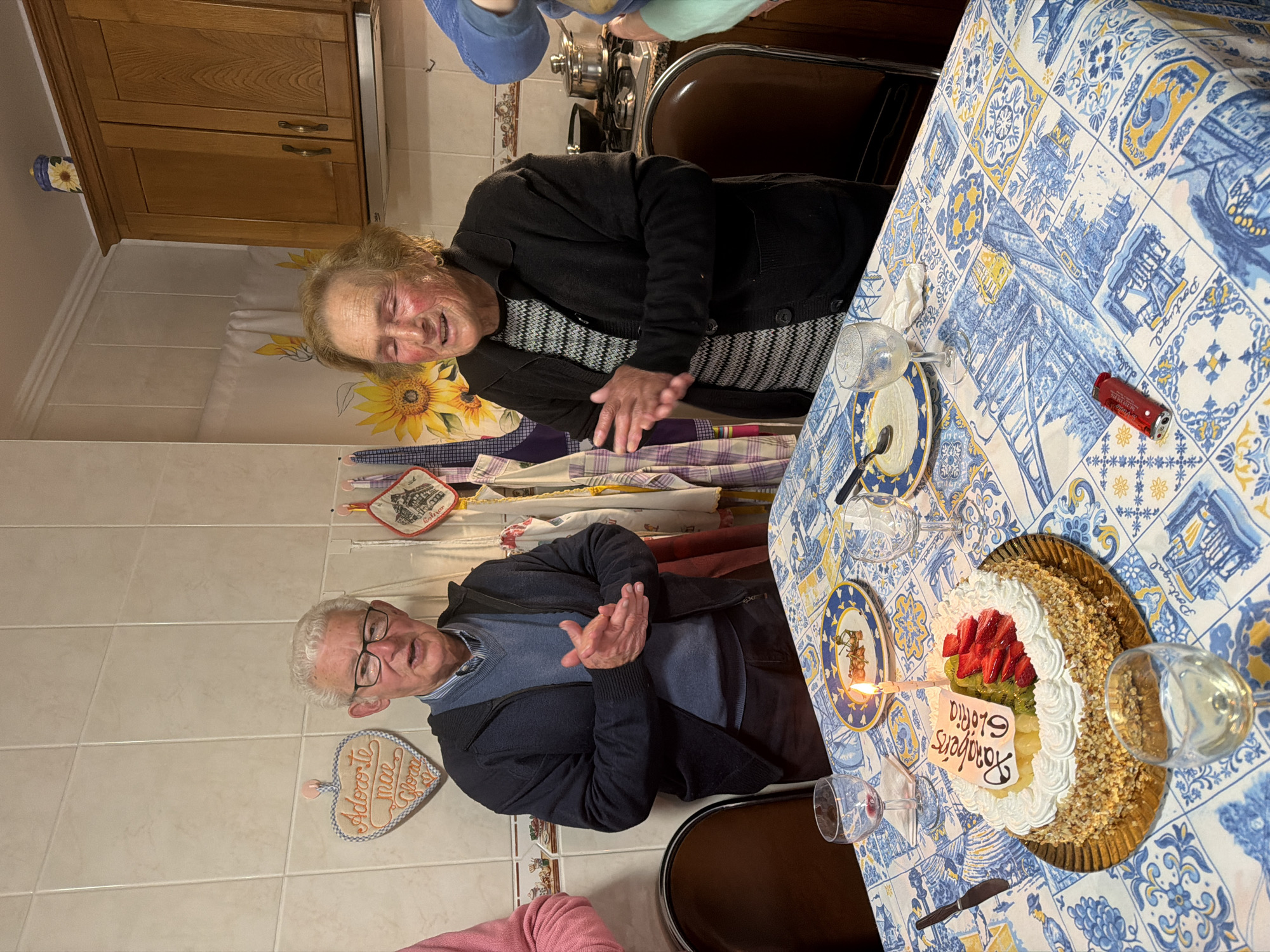 The scene is a birthday celebration for Gloria in Vila Real, Portugal. A man and a woman, likely in their 70s or 80s, are sitting at a table covered with a blue and white Portuguese tile-patterned tablecloth. They are clapping, possibly singing "Happy Birthday". On the table is a birthday cake decorated with strawberries, kiwi, and nuts, and a single lit candle. "Parabens Gloria" (Congratulations Gloria) is written on the cake. There are wine glasses, plates with decorative rims, and cutlery. A red lighter is also on the table. The setting is a kitchen. Wooden cabinets are visible in the background along with a kitchen backsplash decorated with sunflower designs. A heart-shaped wall decoration that reads "Adorote mae Gloria" (Love you Mom Gloria) is also visible. Other people are partially visible behind the two individuals.