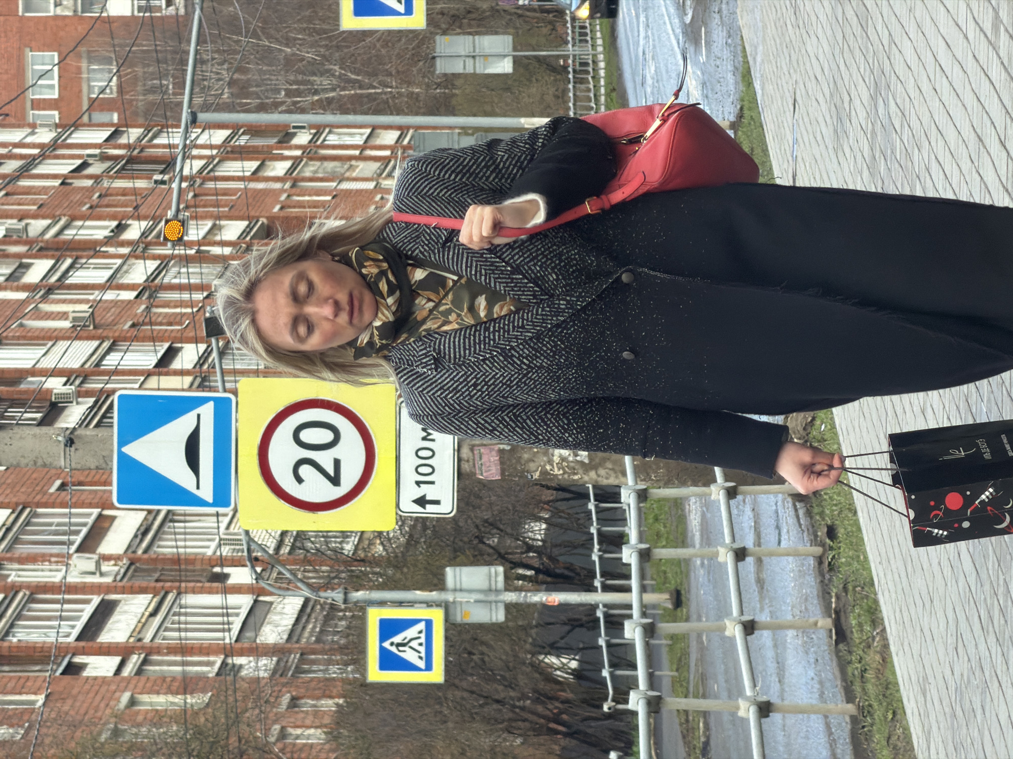 The image captures a woman walking along a city sidewalk in Izhevsk, Russia. She has blonde hair and is wearing a dark patterned coat, a patterned scarf with shades of green and brown, and black trousers. She carries a red shoulder bag over her right shoulder and two black shopping bags in her left hand. Her eyes appear closed or mostly closed, giving her a serene expression.
In the background, there are multi-story residential buildings with numerous windows and balconies. The street features typical Russian road signs, including a blue square sign indicating an uneven road or speed bump, a yellow square sign with a red circle displaying a "20" speed limit, and a white rectangular sign with an upward arrow and "100 M" (meters), likely indicating a distance to an upcoming feature. Another yellow square sign with a blue triangle and pedestrian symbol indicates a pedestrian crossing ahead. A flashing yellow traffic light is also visible. The pavement appears wet in areas, suggesting recent rain. A metal barrier runs along the side of the sidewalk.
The shopping bags she carries have the text "МЭДЖИД ЭКСТЕРИО" (Majid Exterior) along with some decorative patterns featuring planets and rockets. The scene takes place during the daytime, under what appears to be an overcast sky.