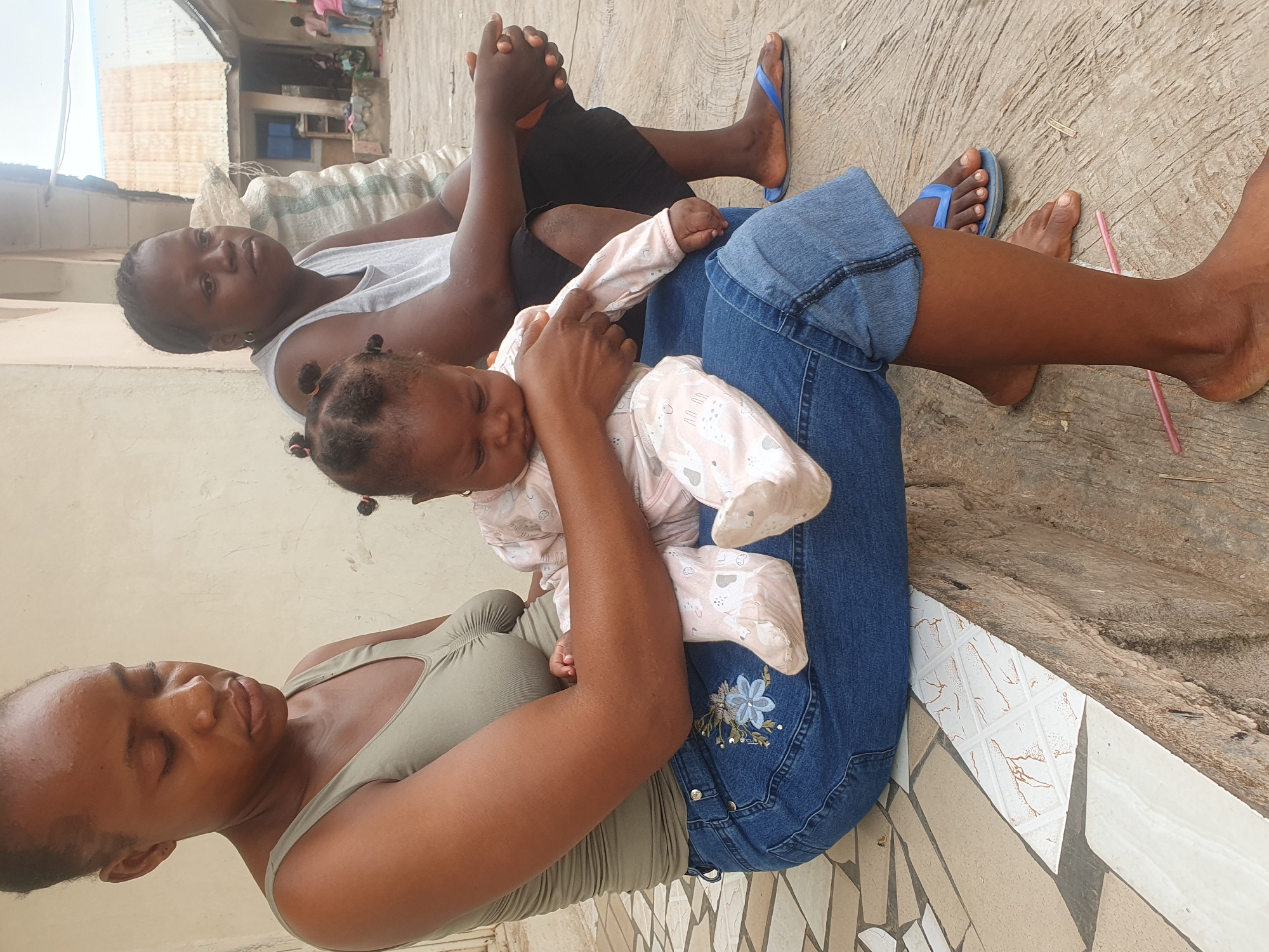 The image shows three individuals sitting outside in Lokoja, Nigeria. A baby wearing a light pink and white patterned pajama set is held by a woman wearing a green tank top and denim capri pants with flower embroidery. Behind them, another person wearing a gray tank top and black shorts is seated. All are barefoot or wearing sandals, sitting on a textured concrete surface next to a tiled area. A pink straw lies on the ground beside the people. Buildings and other individuals are visible in the background.