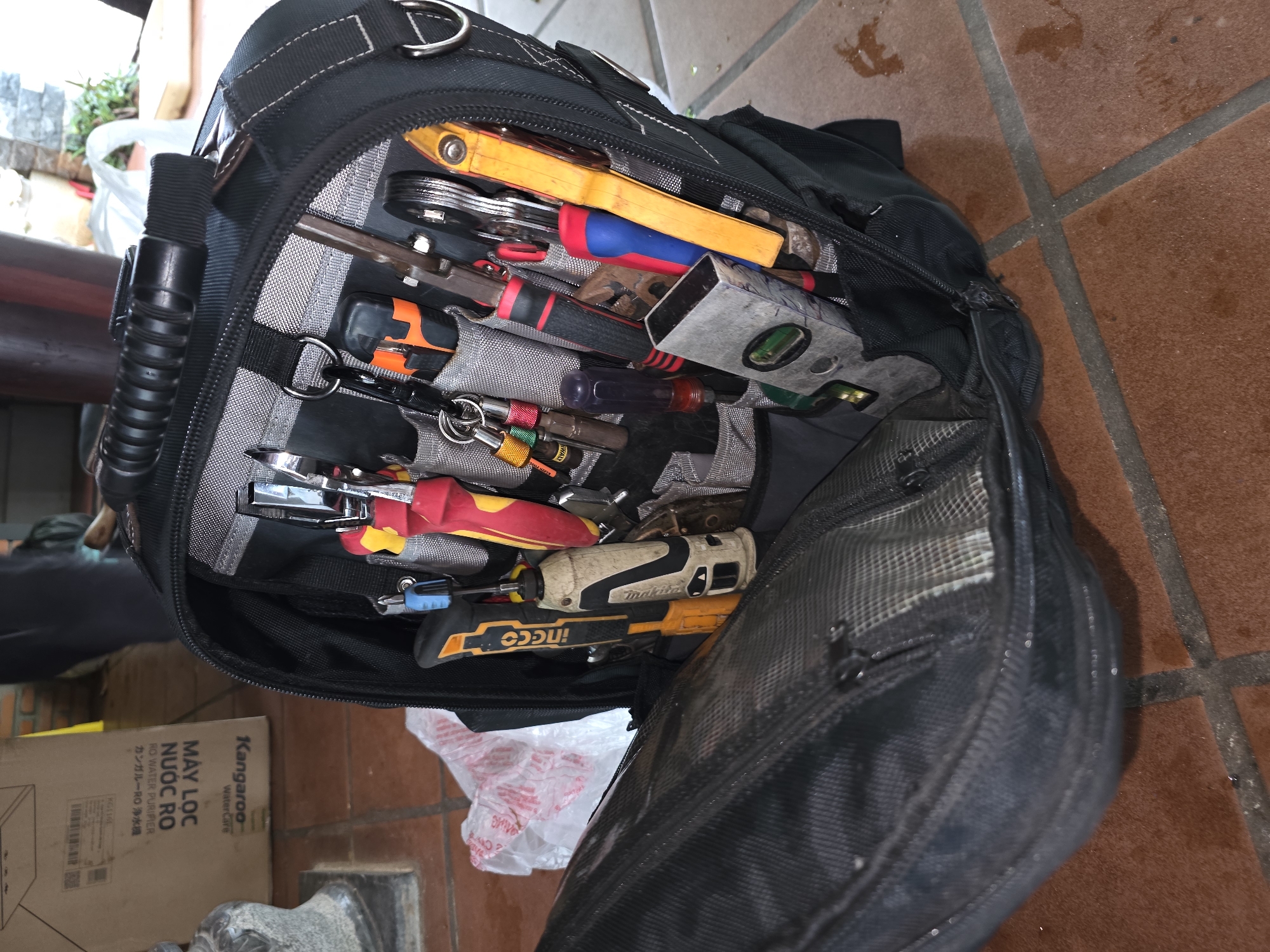 The image shows a black tool bag filled with various tools, including screwdrivers, pliers, and a level. The bag is partially open, revealing its contents arranged in a structured manner. The background includes a cardboard box with Vietnamese text and the logo of Kangaroo WaterCare, suggesting it is a water purifier box. The tools are of different brands and colors. The tool bag sits on what appears to be an orange tile floor, and the presence of a column suggests it's an interior or semi-exterior space. Given the information, the location is most likely in Mũi Né, Vietnam.