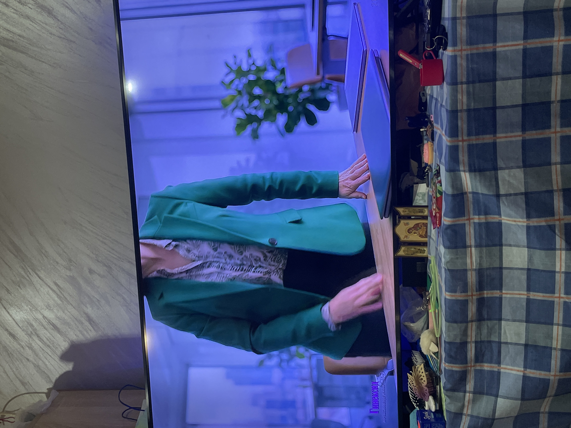 A vertically oriented photograph captures a television screen displaying an interior scene, flanked by elements of the room where the photo was taken.
On the television screen, a person is visible from the waist up, wearing a bright green blazer over a light-colored, possibly animal-print shirt. Their hands rest on a light-toned desk or surface. In the background of the on-screen scene, a large, broad-leafed plant is visible through large windows, suggesting a modern office or professional environment. The entire on-screen image is cast in a prominent cool blue hue, possibly due to lighting or a stylistic filter.
To the right of the television screen, a blue and white checkered blanket or fabric covers a surface, adorned with various small personal items. These include a red miniature backpack or keychain, several other keychains (one black, one light orange/yellow), a small brush with a butterfly design, and what appear to be gold-toned religious icons.
To the left of the screen, a light-colored wall with a textured pattern is visible. A dark shadow, potentially of the photographer, is cast on this wall, and blue cables are present near the bottom edge.
Visible text on the screen includes "Гинексид" in Cyrillic script, located in the bottom right corner (when rotated correctly), with smaller, unreadable text beneath it. The number "21" is also faintly discernible on an object in the upper right of the screen's displayed content. The combination of the modern, office-like scene on the screen and the domestic items flanking the television suggests the photo was taken in a home setting in Ihtiman, Bulgaria, while a program was being viewed.