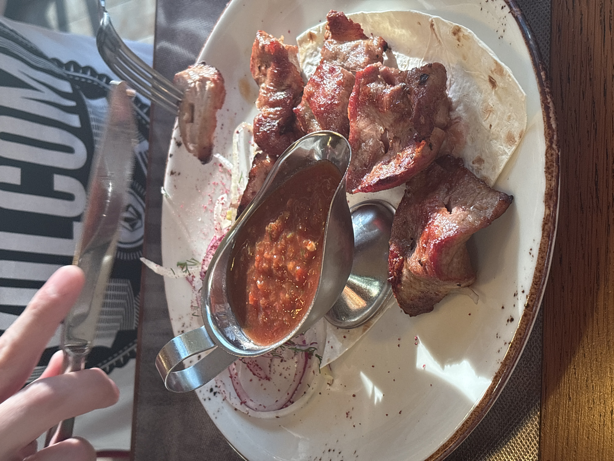 A close-up, high-angle shot shows a plate of meat skewers with a side of sauce and sliced onions. The meat appears to be grilled and is served on a flatbread. The plate is white with a brown rim. A metal gravy boat holds a red, chunky sauce. Sliced red onions are arranged on the plate with some green herbs. In the background, a person's hands are visible, holding a knife and fork, preparing to eat. The person is wearing a black and white striped shirt. The scene is lit by natural light, casting shadows on the plate. The image was taken in Saint Petersburg, Russia.