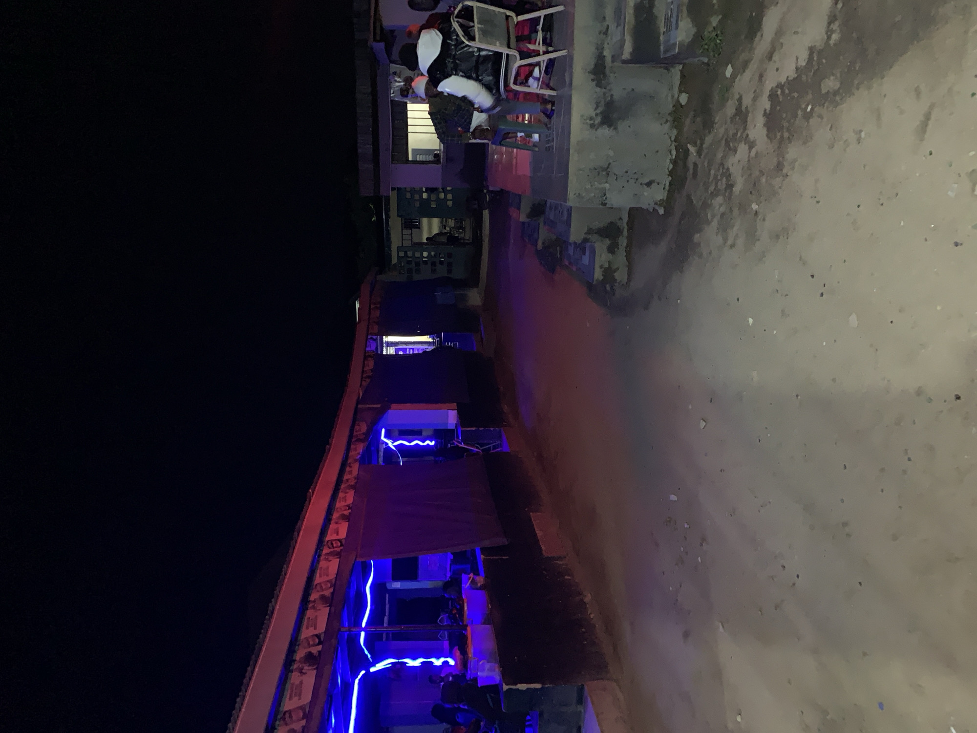 The image depicts an outdoor, nighttime scene in what appears to be a commercial or social establishment in New Stadium, Nigeria. The foreground shows an unpaved, dirt ground scattered with small debris, illuminated by artificial light reflecting from the surrounding structures.
On the left, a single-story building features a red-painted roof fascia. Below the fascia, a banner or frieze displays multiple photographic portraits. The interior of this building is brightly lit with squiggly blue neon strip lights, revealing several individuals seated inside, their forms partially obscured by the lighting and distance. A dark fabric or curtain is drawn across one section of the building's facade.
To the right, a multi-tiered concrete platform or steps leads to another building area. Two individuals are seated on this platform in the mid-ground. One person, wearing a dark puffer jacket with white sleeves and a white hood, is seated on a light-colored metal folding chair, facing left. Another person, partially visible, sits on a green plastic stool nearby. Further back, another structure with a distinctive concrete block screen design and a window is visible, with faint figures present within. The overall sky is dark.