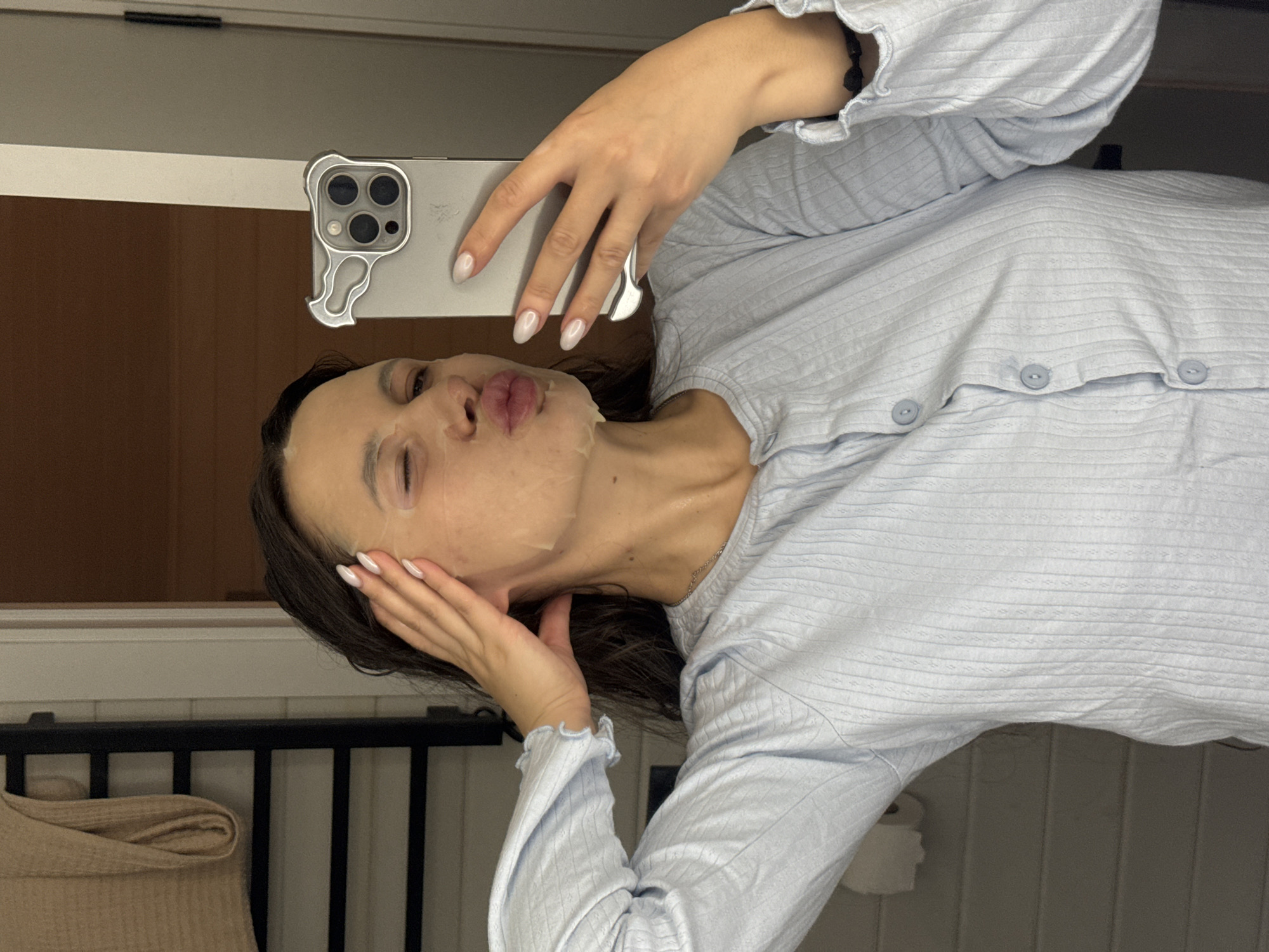 A woman is captured in a mirror selfie, posing playfully with puckered lips while wearing a translucent sheet mask on her face. She has long, dark hair and light-colored, almond-shaped manicured nails. She is dressed in a light blue, ribbed long-sleeved top featuring ruffled cuffs and three visible light blue buttons down the front, suggesting loungewear or pajamas. One hand holds a silver-colored smartphone with a distinctive case that outlines the camera module, while the other hand rests near her cheek. A thin silver necklace is also visible.
The setting appears to be indoors, likely a private residence in Литвиново, Russia, given the personal nature of the activity. The background includes a dark metal railing, possibly part of a bed frame or room divider, and a folded beige textile object. A roll of white paper, likely toilet paper, is partially visible in the background, suggesting the location could be a bathroom or bedroom. The activity indicates a relaxed moment of self-care, likely taking place in the evening or at night. There is no discernible text in the image.