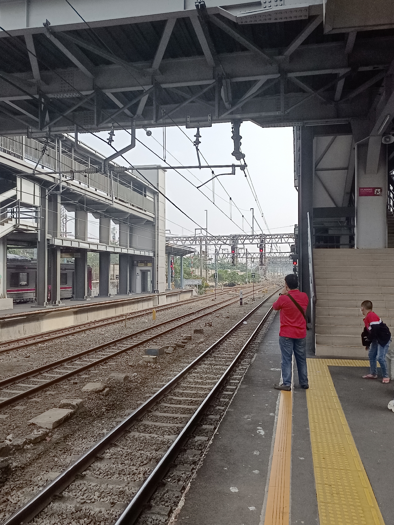 A man in a red shirt and blue jeans stands on a train platform in Central Jakarta, Indonesia, facing away from the camera and holding a camera or phone to his eye, appearing to take a photograph of the train tracks. Next to him, a child in a black jacket with red trim and blue jeans stands on the platform, looking towards the tracks. A yellow tactile paving strip runs along the edge of the platform. In the background, train tracks stretch into the distance, with a train visible on the left side of the image. Overhead, a large, complex network of metal beams and wires supports the station's infrastructure. A set of stairs leads up to a higher level of the station. A red sign with white text and a symbol is visible on a pillar near the stairs. The sky is overcast.