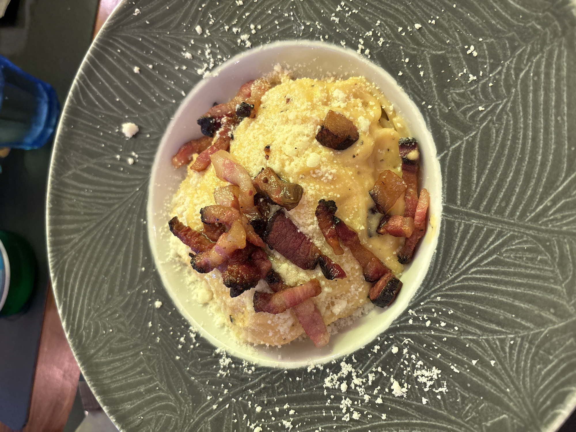 A close-up, top-down shot of a white bowl filled with pasta topped with crispy bacon and grated cheese, presented on a dark gray patterned plate. The pasta appears to be coated in a creamy yellow sauce. Scattered white specks, likely cheese or salt, dust the pasta and the plate's rim. The background is blurred, hinting at a restaurant setting with a dark wooden table and possibly drinks in blue and green bottles. The specific location in Barcelona, Spain, is not discernible from the image itself.