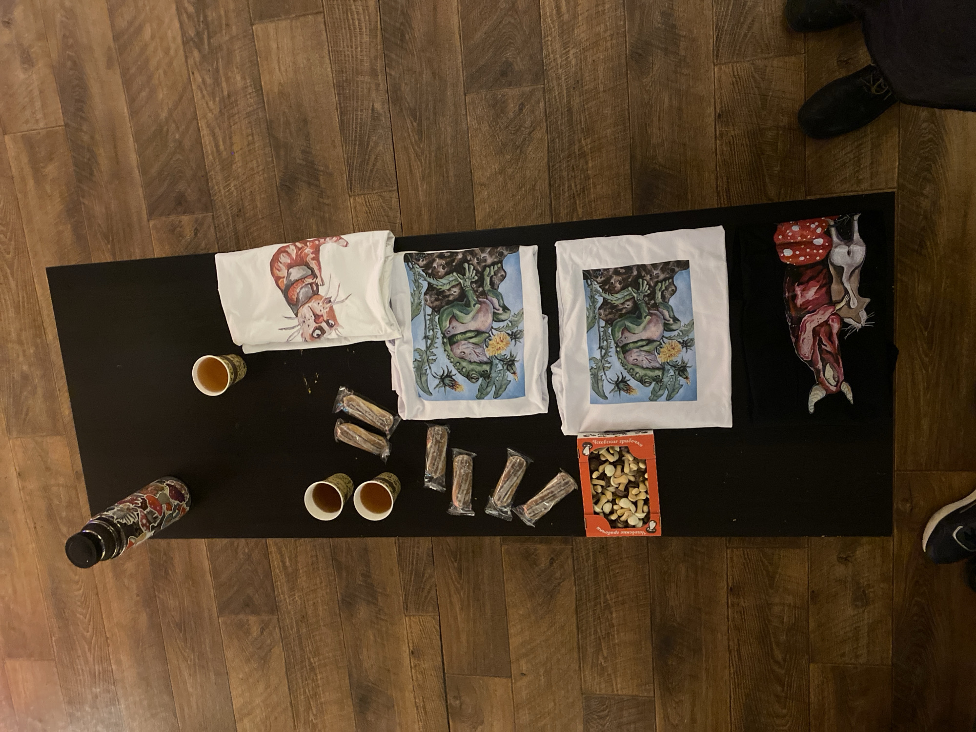 The scene presents an overhead view of a dark, rectangular surface, likely a table or mat, positioned on a textured, wide-plank wooden floor in Moscow, Russia.
**Objects:**
A sticker-adorned water bottle with a black cap rests diagonally in the upper-left of the dark surface. Three small paper cups, two grouped on the left and one on the right, contain a dark liquid. Six individually plastic-wrapped rectangular snack bars are arranged in two vertical columns of three. A red cardboard box with a transparent lid holds an assortment of nuts and mushroom-shaped confectionery; the box features Russian text including "Лесовые грибочки" (Forest mushrooms). Four folded textile items, consistent with t-shirts, are laid out vertically. The topmost is white with a graphic of a stylized reddish-brown feline-like creature. Below it, two identical white t-shirts feature a graphic of a green winged insect, possibly a dragonfly, amidst dandelions. The bottommost item is a black t-shirt displaying a graphic of an animal with exposed internal organs next to a red mushroom with white spots.
**People and Environment:**
No complete individuals are visible. Portions of two feet are present on the wooden floor: one wearing a dark blue and white sneaker in the lower-left corner, and another wearing a black shoe in the lower-right corner, indicating the presence of at least two persons standing around the display. The wooden floor planks exhibit a pronounced grain and distressed appearance. The overall arrangement suggests a casual display of merchandise and refreshments.