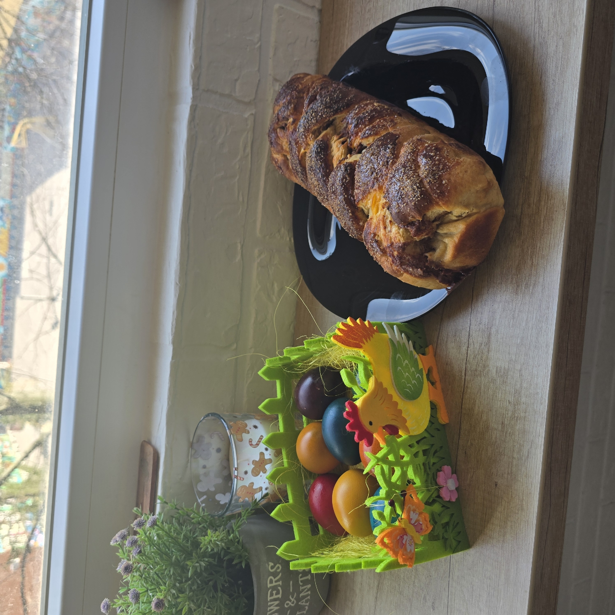 On a table in front of a window, likely indoors, is a braided bread on a black plate, and an Easter decoration containing colored eggs, a yellow chicken, and orange butterflies. The scene could be located in Sofia, Bulgaria. A pot with a plant is also visible.