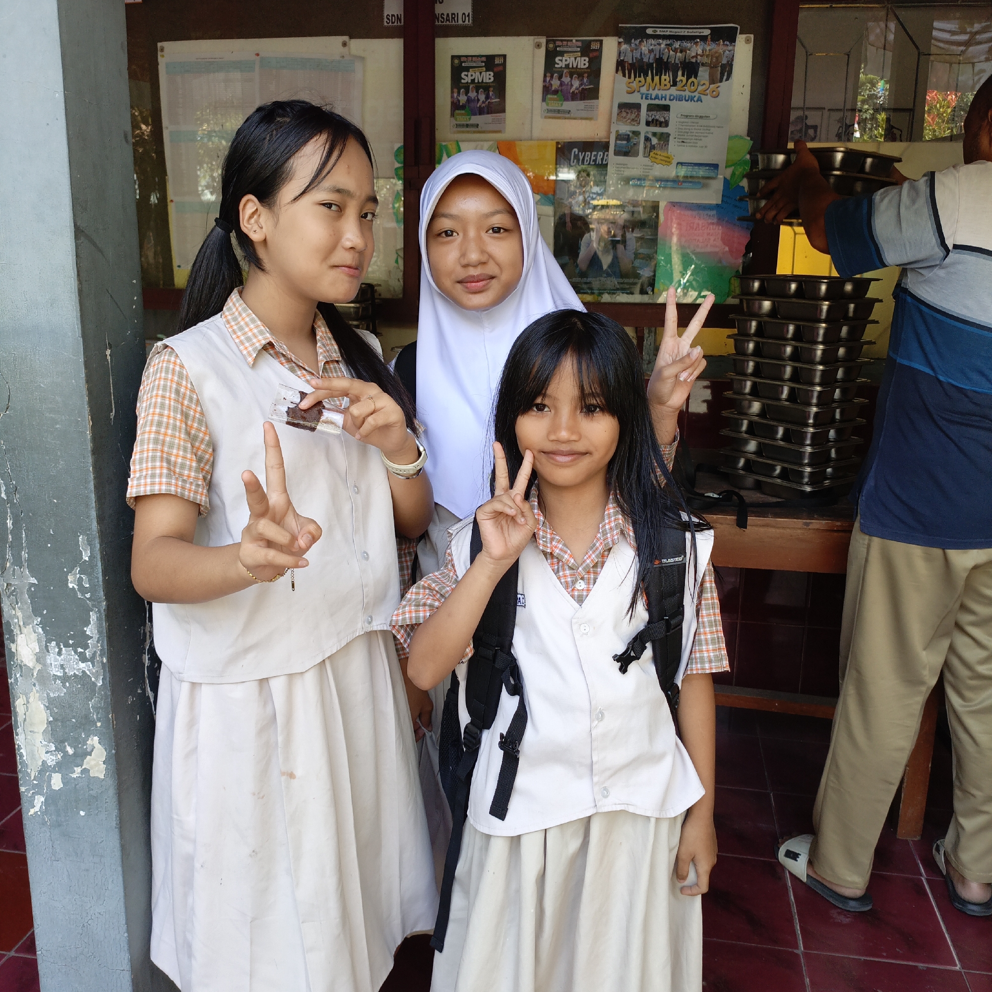 Three young females, appearing to be students in uniform, pose for a photograph. The student on the left, with her hair in pigtails, holds a small transparent packet containing dark brown food and makes a peace sign with her right hand. The student in the center wears a white hijab. The student on the right, facing the camera directly, also makes a peace sign with her right hand. All three students are wearing white vests over checkered shirts and light-colored skirts. Behind them, a stack of dark metal trays is visible, and to the far right, a man wearing a blue and white striped shirt and beige pants is partially visible, appearing to be handling the trays. The background includes a bulletin board with various posters, one of which advertises "SPMB 2026". The setting appears to be an outdoor area, possibly a school cafeteria or administrative building, in Salatiga, Indonesia, suggested by the Indonesian text on the bulletin board and the visual style of the architecture. The ground is tiled with red squares, and a rough-textured gray pillar is to the left of the frame.