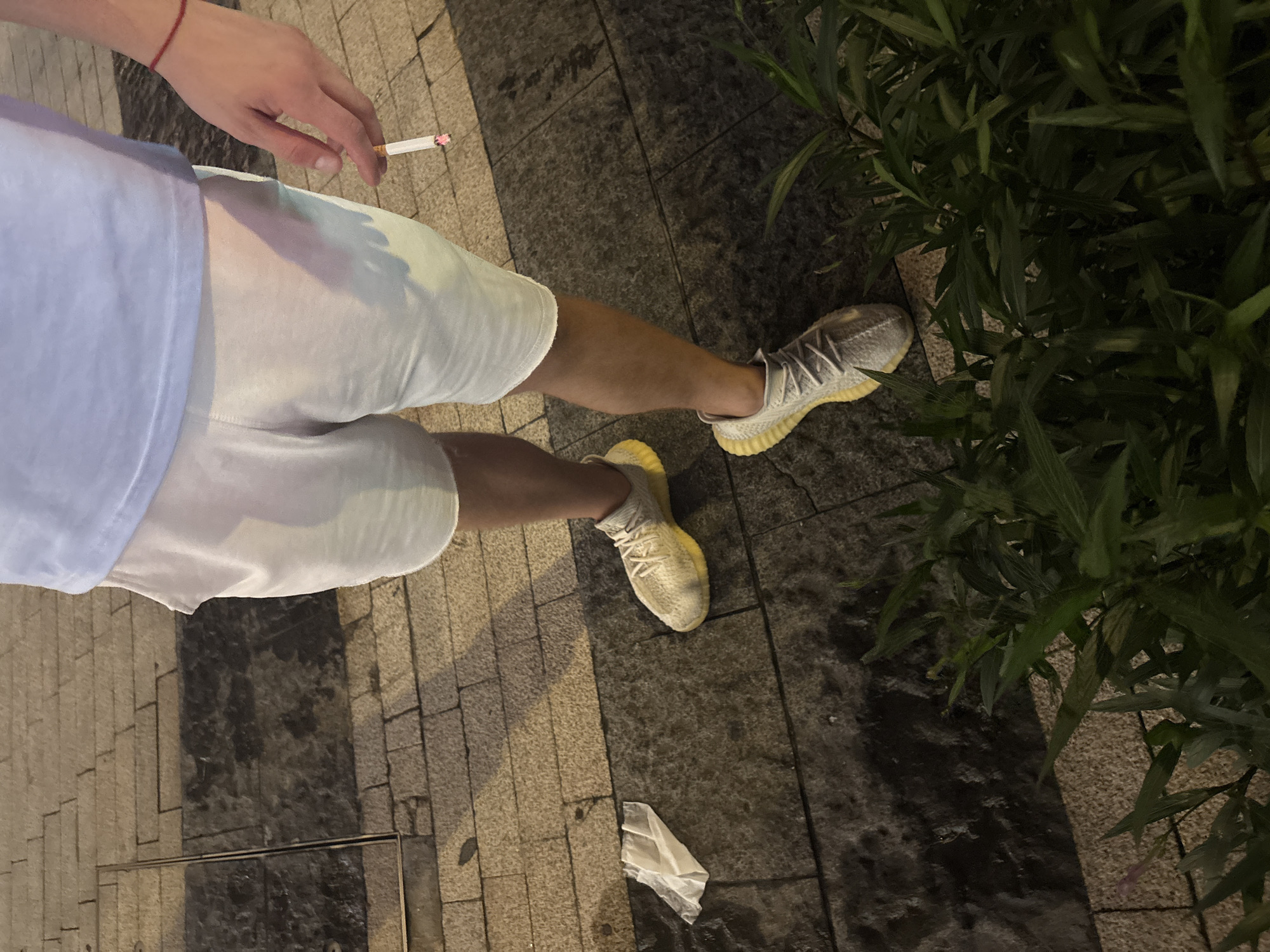 This is a high-angle, full-body shot of a person standing on a sidewalk at night. The person is wearing white shorts and a light blue t-shirt. They are holding a lit cigarette in their right hand. Their feet are clad in off-white sneakers with yellow soles. The sidewalk is paved with light-colored rectangular stones, with darker, possibly wet, patches indicating the time of day or recent weather. To the right of the person, there is dense green foliage, suggesting a park or landscaped area. A crumpled piece of white paper or plastic is visible on the ground near the person's feet. The lighting suggests it is evening or nighttime, with artificial light creating shadows on the ground. The overall mood is casual, with no overt emotional expression visible. No text is discernible in the image. The location context is Tianhe District, China.