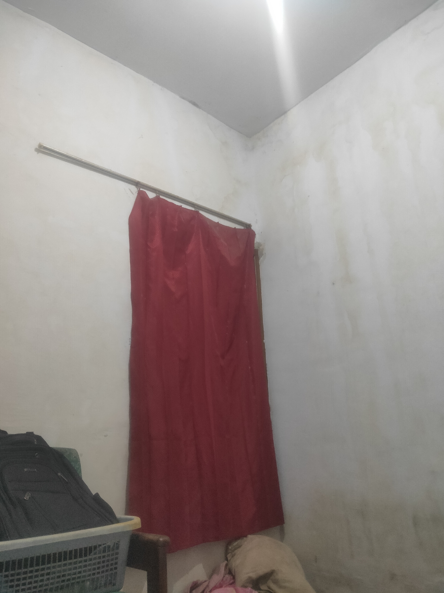 The image shows an interior scene, likely a room, with white walls and a white ceiling. A red curtain hangs from a metal rod, partially obscuring a doorway or window. In the lower left corner, a black backpack is placed in a grey plastic basket. The walls appear somewhat stained or discolored. The scene is lit from above, with a bright light source visible at the top of the image. The overall impression is of a simple, perhaps sparsely furnished, domestic interior. There are no people visible in the image. The location is identified as Antirogo, Indonesia.