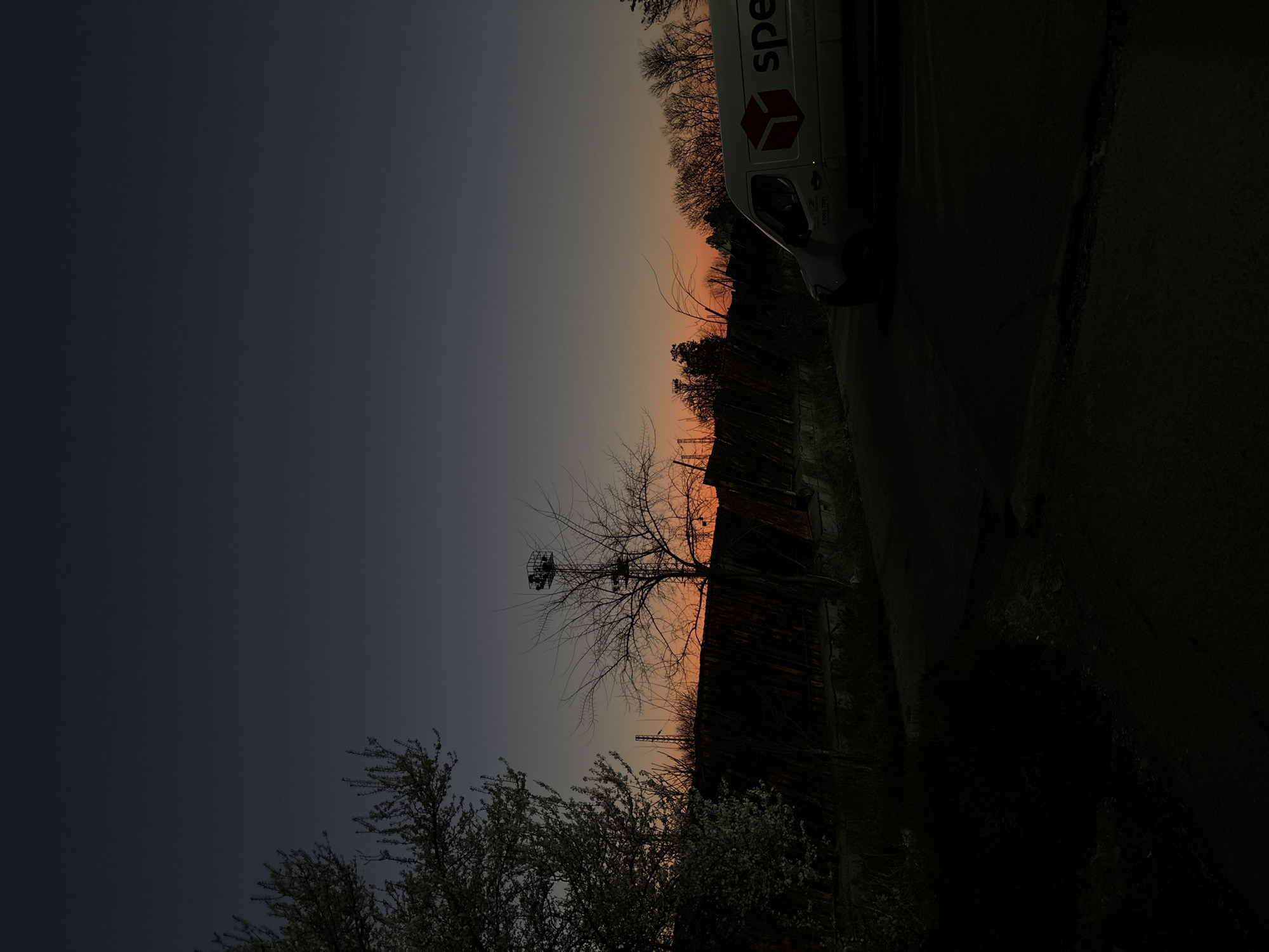 The image presents a dimly lit, dusk scene, likely taken near Burgas, Bulgaria. The sky displays a gradient of colors, transitioning from dark blue-grey in the upper left to orange-red near the horizon. Silhouetted against the sunset are a variety of objects, including bare trees and blooming bushes in the lower left of the frame. A utility pole or tower is positioned centrally in the image. A fence or building runs horizontally across the center of the image. In the upper right portion of the picture, a white van with red lettering is positioned along the side of a road. The location is suggested by the delivery service vehicle visible in the frame, combined with the architectural and environmental elements depicted.