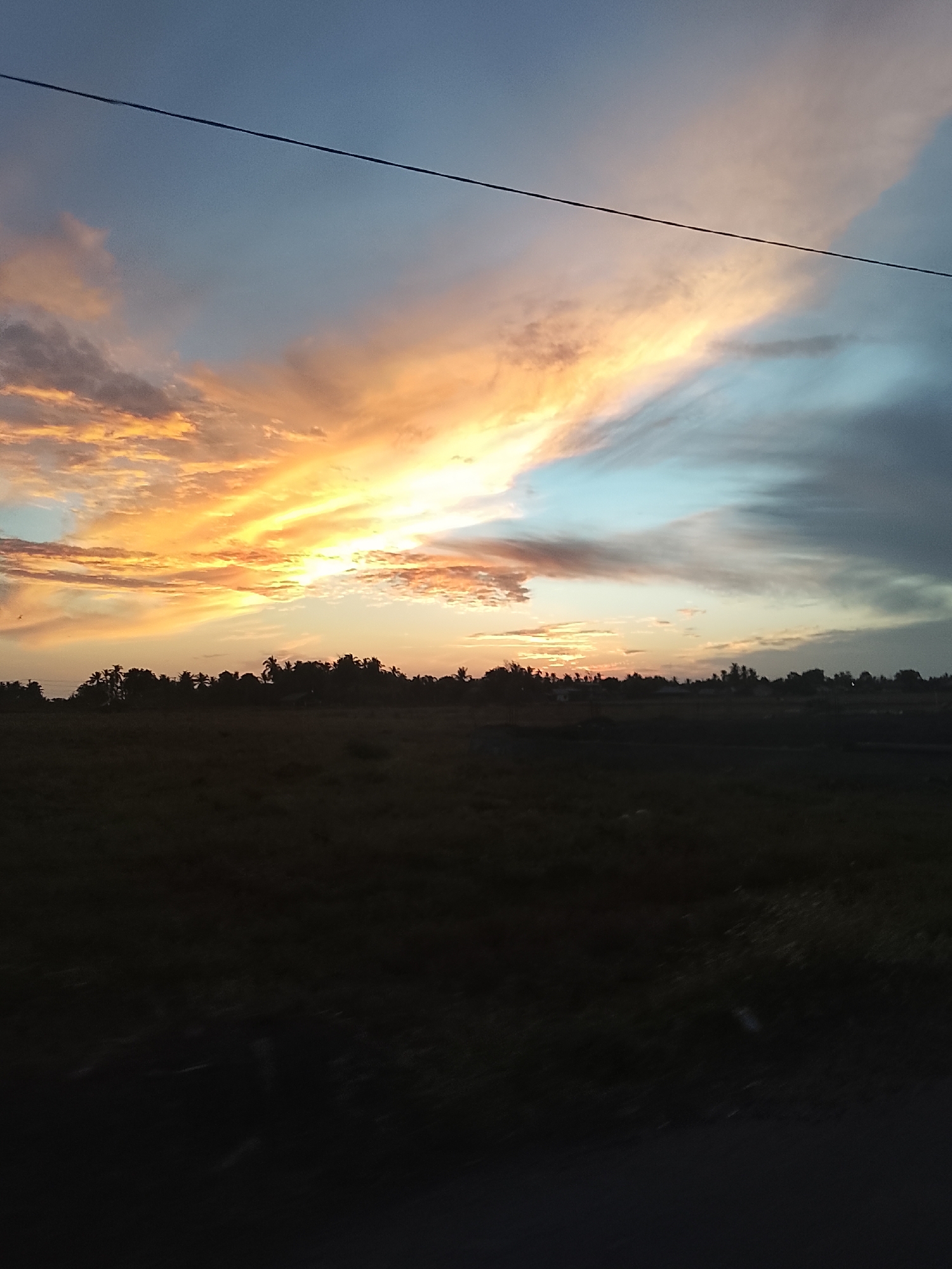 The image depicts a sunset over a rural landscape in Meunasah Jurong, Indonesia. The sky is a dramatic display of orange, yellow, and blue clouds, streaked by a thin, dark wire that traverses the upper portion of the frame. Below the vibrant sky, a dark silhouette of trees forms a tree line, with hints of buildings or structures visible within the foliage. In the foreground, a dark, out-of-focus field stretches across the bottom of the image, suggesting a natural or agricultural area. The overall scene is one of natural beauty and tranquility, with the setting sun casting a warm glow.