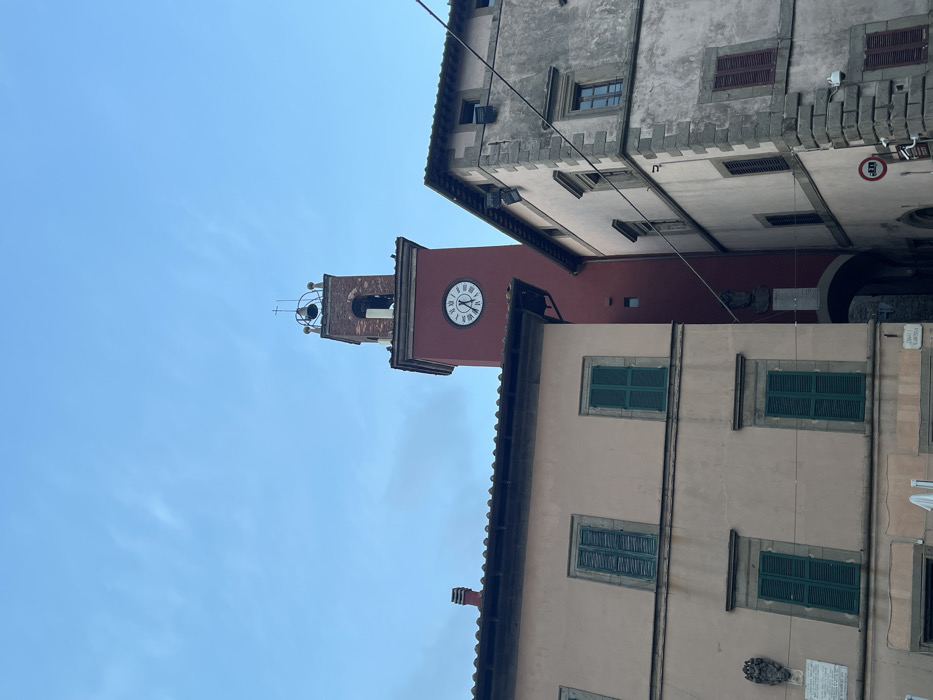 An upward-angled view captures a historic street scene in Castel del Piano, Italy, dominated by a prominent reddish-brown clock tower situated centrally. The tower features a white clock face with black Roman numerals, indicating approximately 1:55 or 2:55, and is topped with a belfry containing a bell and a small cross. An archway at the base of the tower leads into a shadowed passage.
To the left, a two-story building with a light beige facade presents four windows, each equipped with closed, dark green louvered shutters. A decorative crest and a white plaque are affixed to the wall on the lower left. A chimney stack is visible above its tiled roofline.
To the right, a multi-story building with a weathered, light grey facade displays stone quoins on its corners. Several windows, some with closed dark red shutters, are visible. Utility lines span horizontally across the upper portion of this building and the space towards the clock tower. A circular red and white 'no entry' sign, accompanied by a smaller rectangular sign, is mounted on the lower right corner of this building. Outdoor lighting or security cameras are also visible on its upper facade. The sky above is a light, clear blue with minimal cloud cover. No individuals are present in the visible scene.