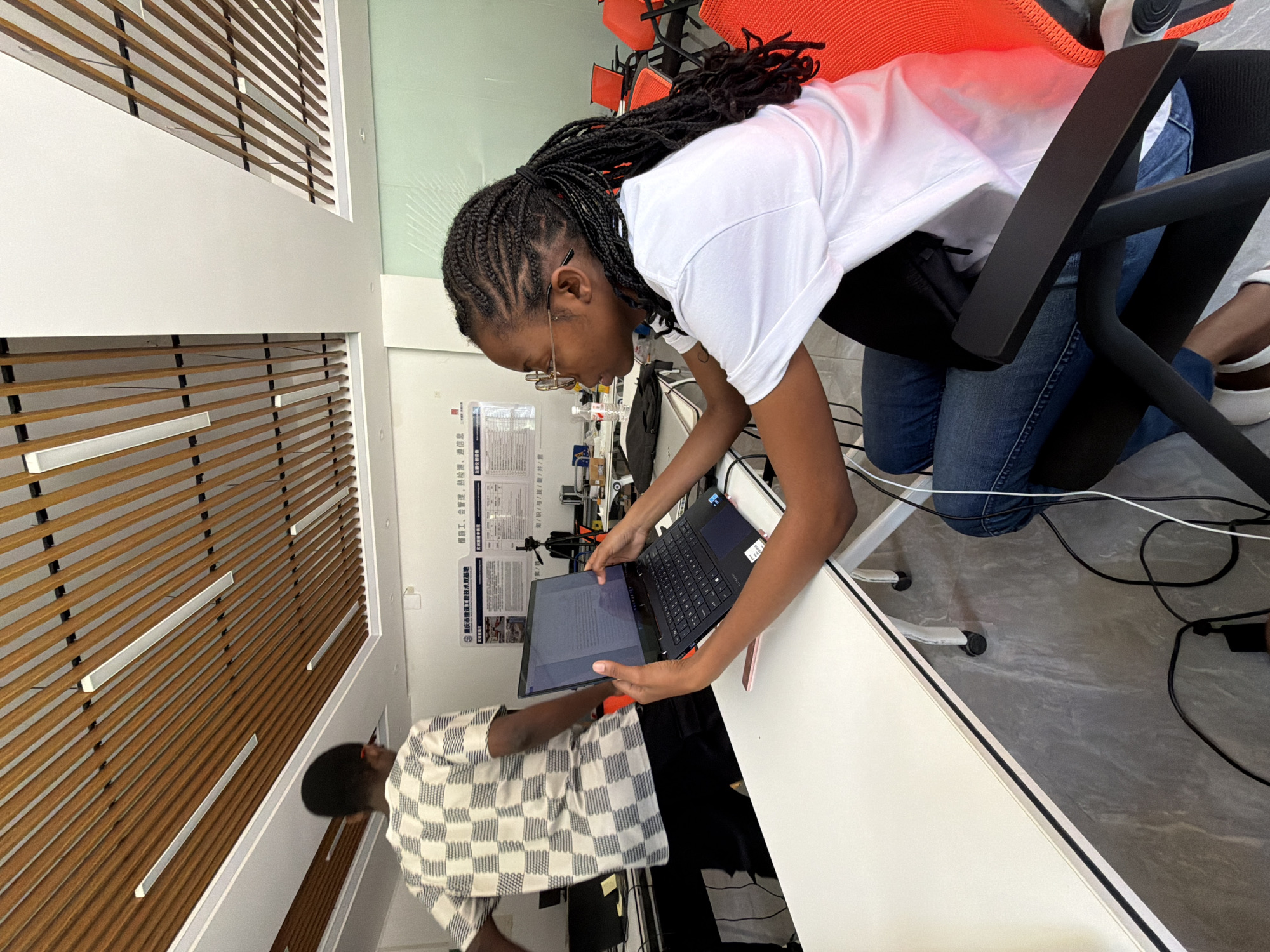 In the city of Jiangjin, China, a young Black woman wearing glasses and a white t-shirt with braided hair sits at a white desk, looking at a laptop. She is wearing blue jeans and white shoes. Another person, a Black male wearing a checkered shirt and dark pants, is walking in the background. The room appears to be an office or classroom with a modern ceiling featuring wooden slats and strip lighting. There are orange chairs visible in the background, along with a whiteboard or poster on the wall with Chinese writing. A water bottle and other desk accessories are present. Cables are visible on the floor.