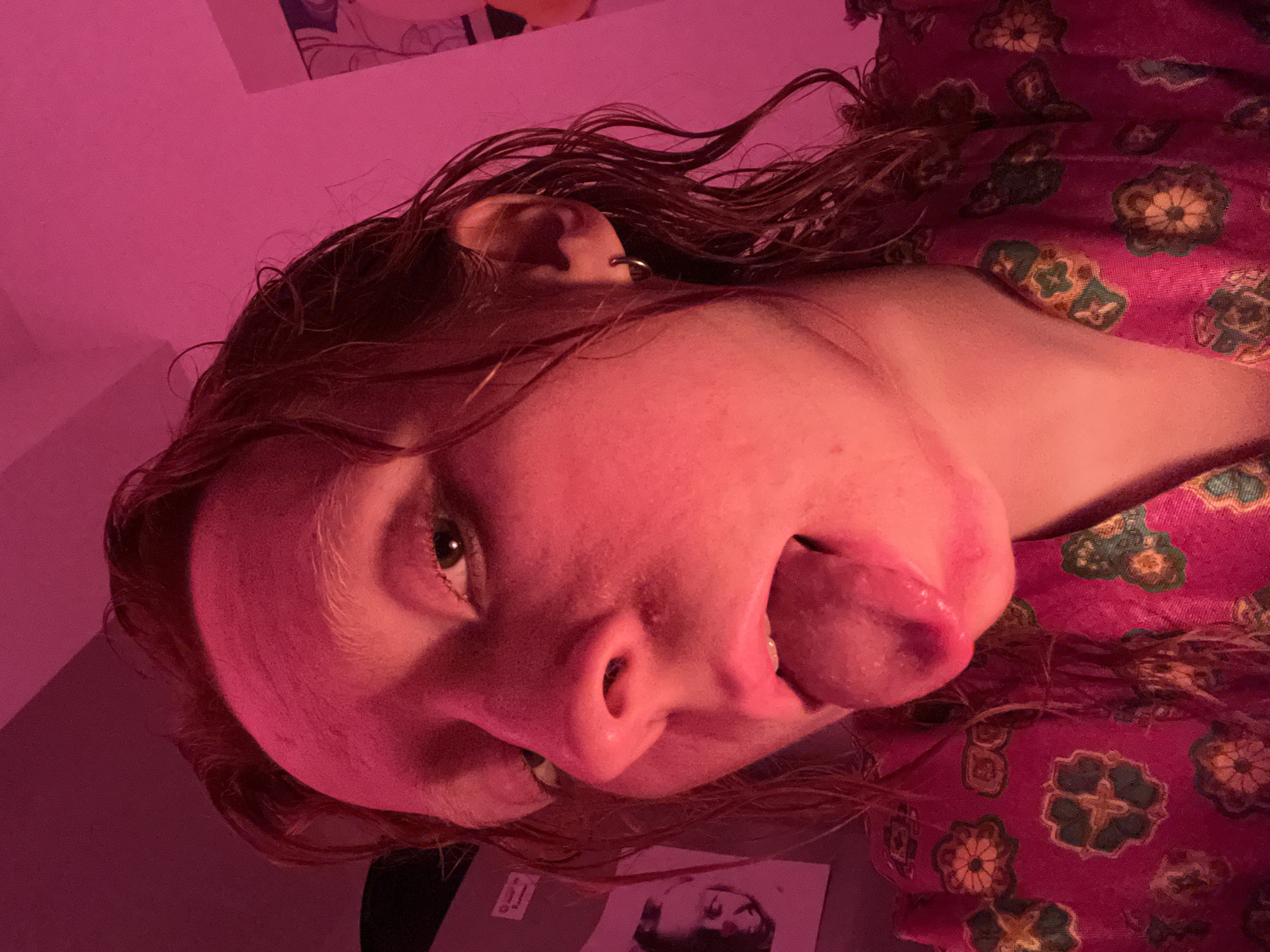 A fair-skinned person with reddish-brown hair is lying on their side, sticking their tongue out. They have an earring in their left ear. They are wearing a pink and green patterned shirt. The lighting is pink. A poster can be seen behind the person's head. Another image is visible near their lower left side. The location is impossible to determine with the information given.