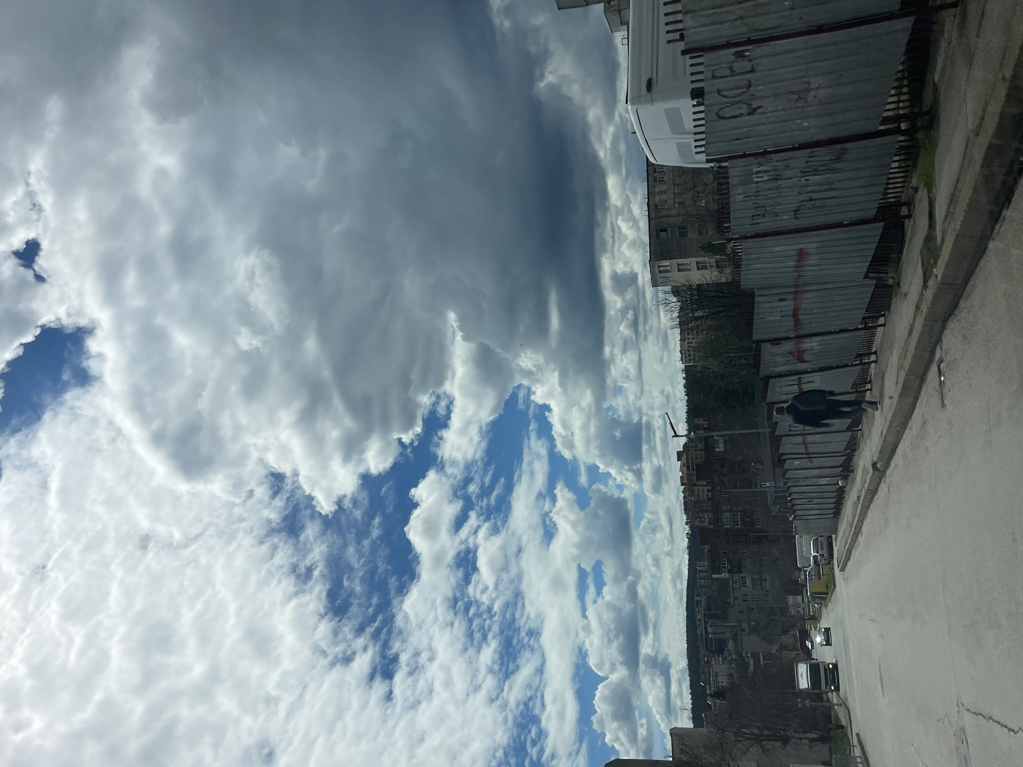 The image shows a street scene in Veliko Tarnovo, Bulgaria, under a cloudy sky. To the right, a corrugated metal fence runs along the edge of the sidewalk. A person walks along the sidewalk beside the fence. Behind the fence, buildings of varying heights form the urban landscape. The sky is a mix of blue patches and dense white and gray clouds. Cars can be seen parked and driving along the street.