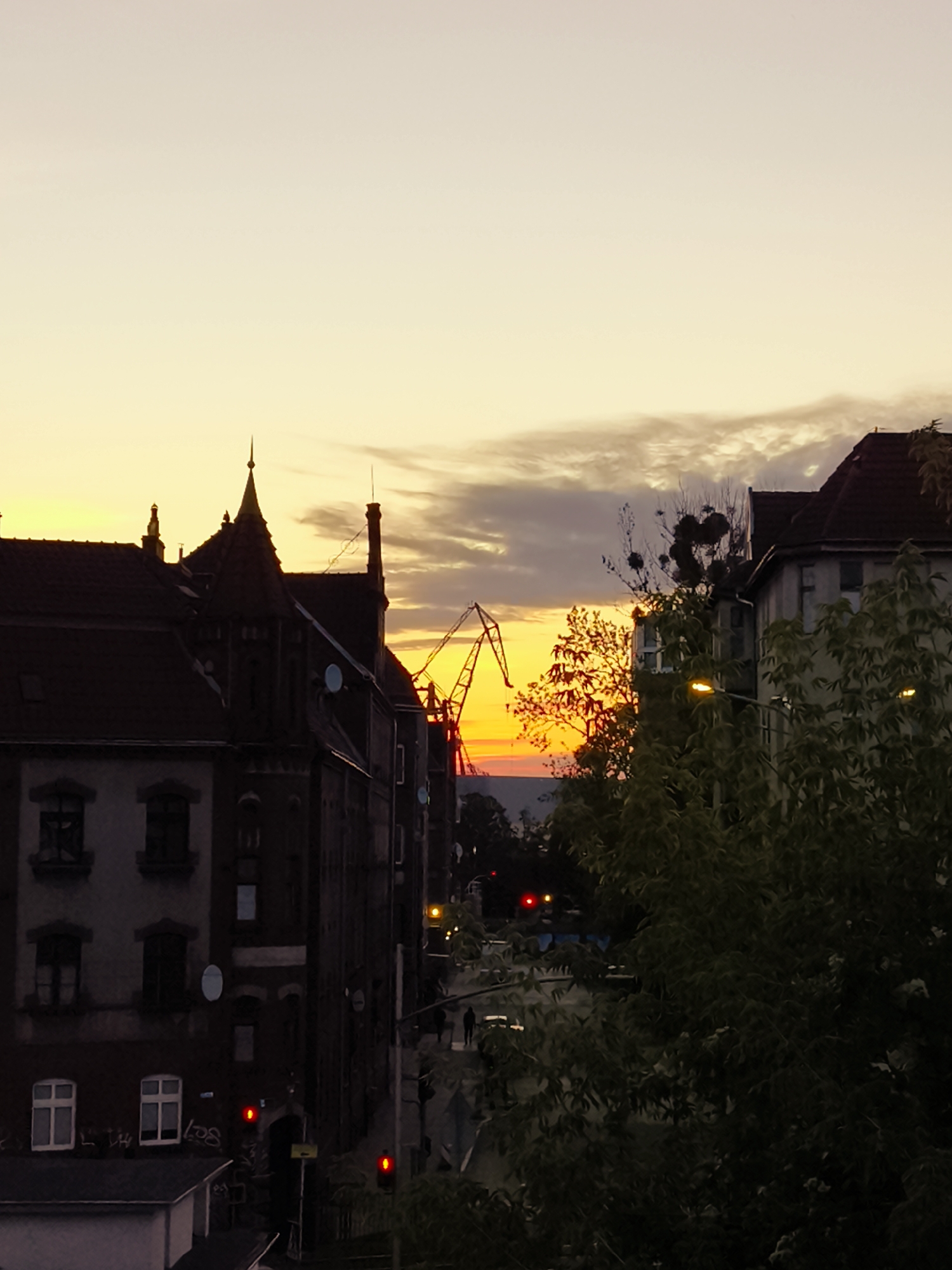 The image shows a sunset over the city of Gdańsk, Poland. In the foreground, there are several buildings with dark red roofs. One building on the left has a distinctive pointed turret. To the right, a large green tree partially obscures a building. In the background, a yellow crane is visible against the orange and yellow hues of the sunset. The sky is a gradient of pale yellow at the horizon, transitioning to a lighter shade upwards. There are some clouds scattered across the sky. The street level shows traffic lights, and silhouetted figures of people walking.