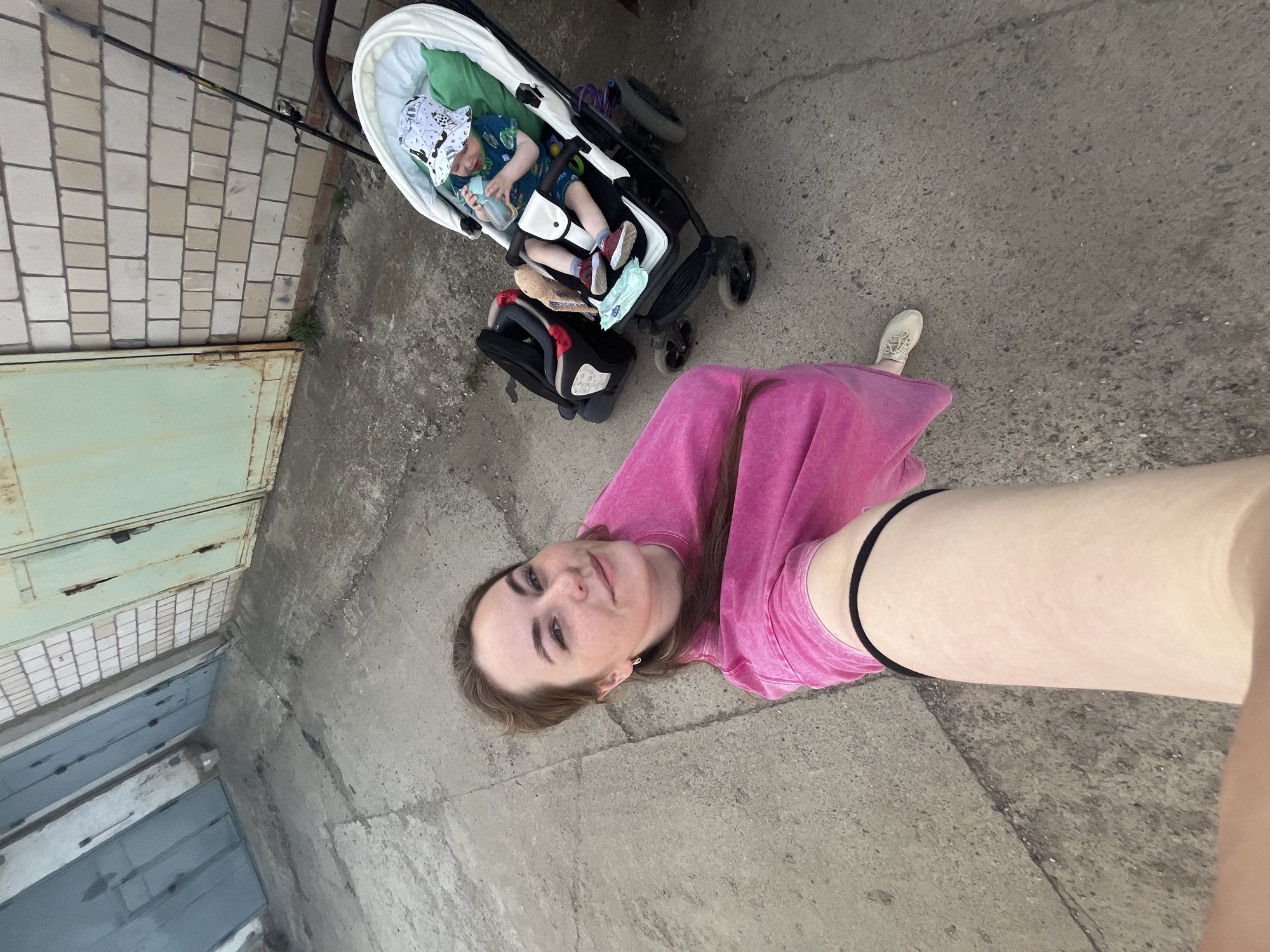 A selfie taken outdoors in what appears to be an urban alleyway or service area in Chaikovsky, Russia. A woman is visible from the waist up, wearing a pink top. A baby is in a stroller with a white interior; the baby is holding a bottle. A black car seat is placed next to the stroller. The environment features a concrete ground and wall, with metal garage doors visible in the background. The weather appears to be overcast.