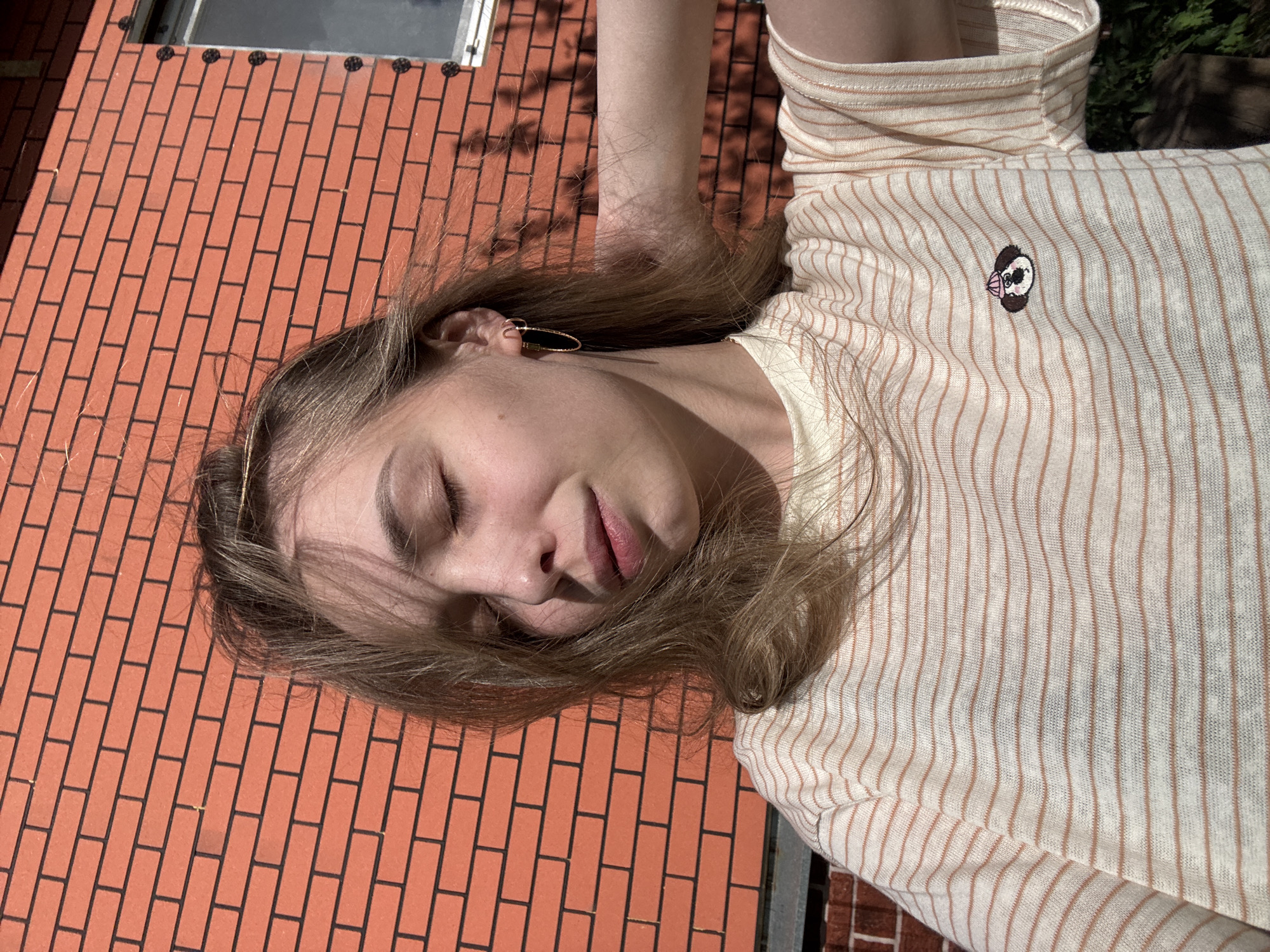 A woman with long blond hair is lying on a brick surface, possibly a patio or rooftop. She is wearing a cream-colored turtleneck sweater with brown stripes and a small embroidered dog on the chest. Her eyes are closed. The scene is outdoors, and a window or skylight is visible in the background. The location is indicated to be Кемуль, Russia, but there is no indication within the image that confirms the location.