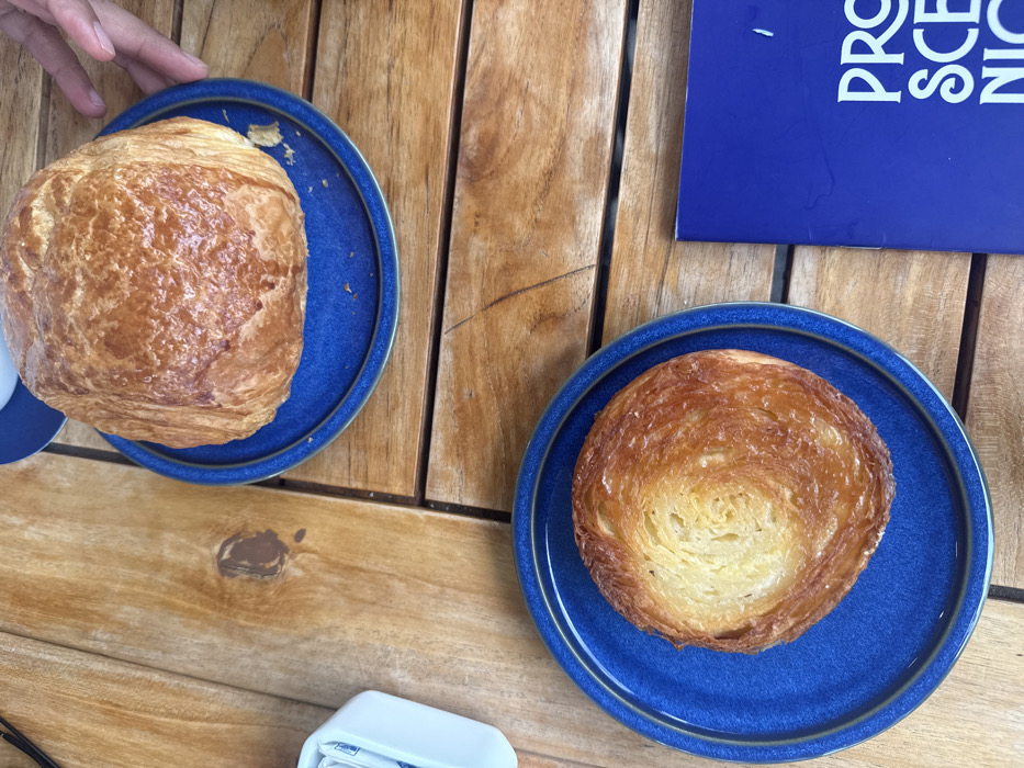 An overhead, high-angle shot depicts a tabletop scene in a cafe or restaurant in Mexico City, Mexico. The table surface consists of light brown wooden slats. Two distinct pastries are presented on circular, deep blue ceramic plates. The upper plate, positioned towards the top-center, holds a large, rounded, golden-brown baked good with a visibly flaky exterior. A human hand, displaying light skin tone, is partially visible on the right side of this upper plate, with fingers resting near its rim. The lower plate, positioned towards the bottom-center, contains a flatter, circular pastry characterized by a prominent spiral pattern of golden-brown, layered dough. To the upper left, a white rectangular container holds multiple small, white-wrapped packets, consistent with sugar or sweetener. In the lower right quadrant, a portion of a blue object, identified as a book or menu, is visible, displaying white sans-serif text that begins "PRO SCEN NIC...". The lighting suggests an outdoor or brightly lit indoor environment.