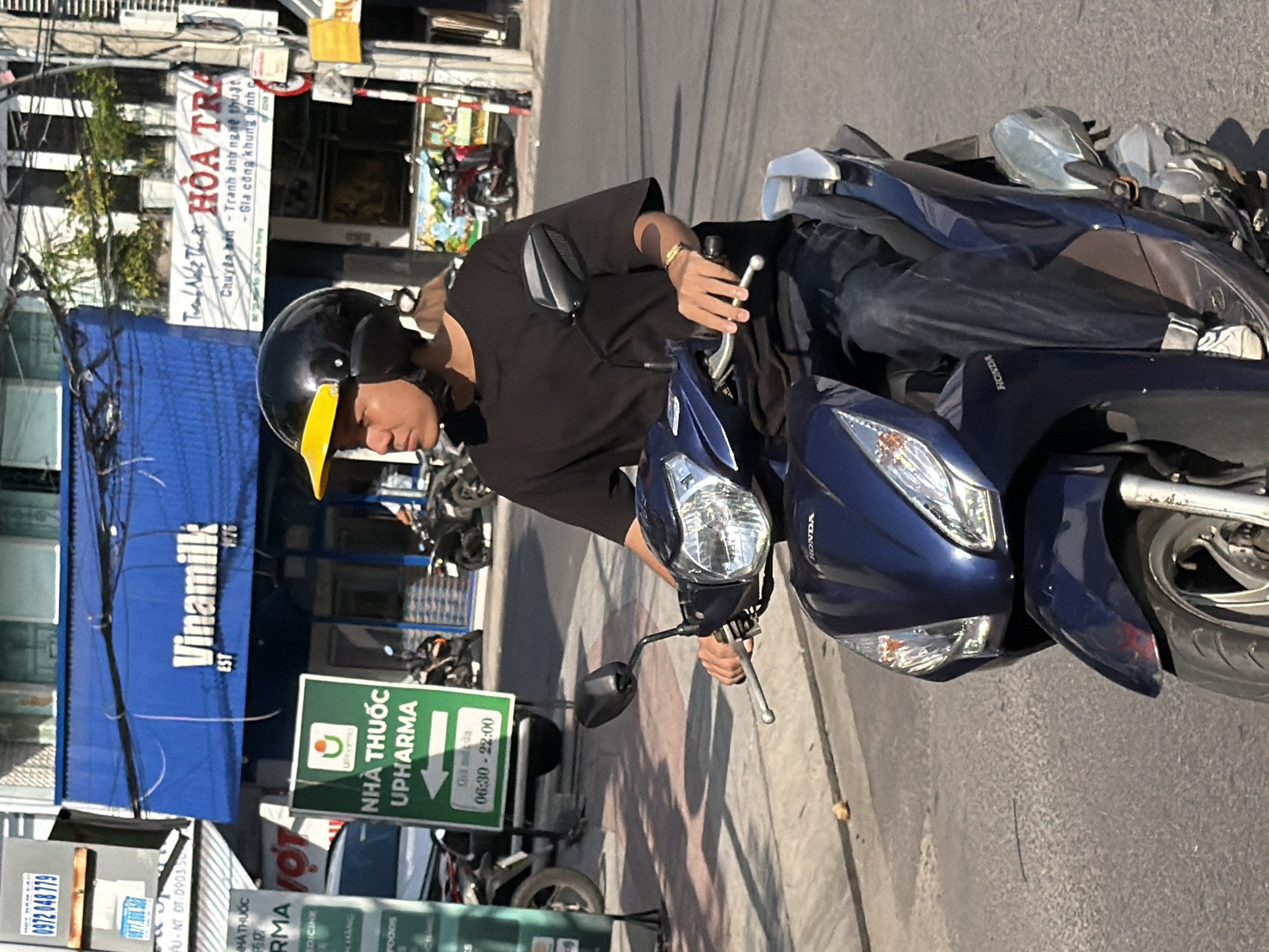 A medium shot captures a young man wearing a black helmet with a yellow brim and a black t-shirt, riding a dark blue Honda scooter on a city street in Nha Trang, Vietnam. The man's left hand is on the scooter's handlebars, and he is looking to his left. Behind him, a building with a bright blue awning displays the "Vinamilk" logo, a well-known Vietnamese dairy company. To the left of Vinamilk, a green sign indicates "NHÀ THUỐC UPHARMA," a pharmacy, with operating hours listed as 06:30 - 22:00. The street appears to be paved, and there are other motorcycles parked in the background, along with a white car and the facades of various businesses. The lighting suggests it is daytime, with shadows cast on the ground.