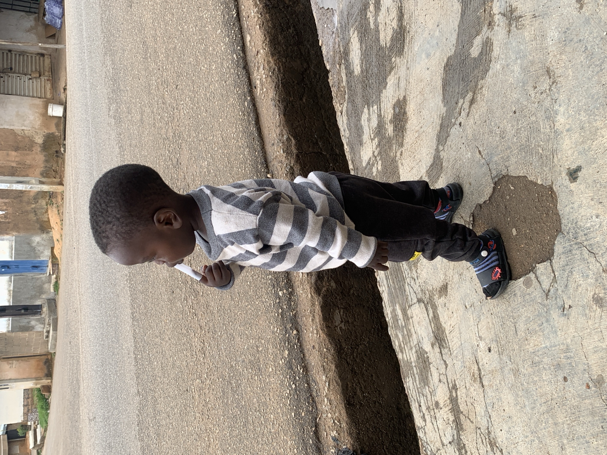 A young child stands on a concrete sidewalk in Gura Top, Nigeria, adjacent to a paved street. The child wears a grey and white striped sweater, dark trousers, and black sandals. They hold a small, white object near their mouth. Buildings are visible in the background.