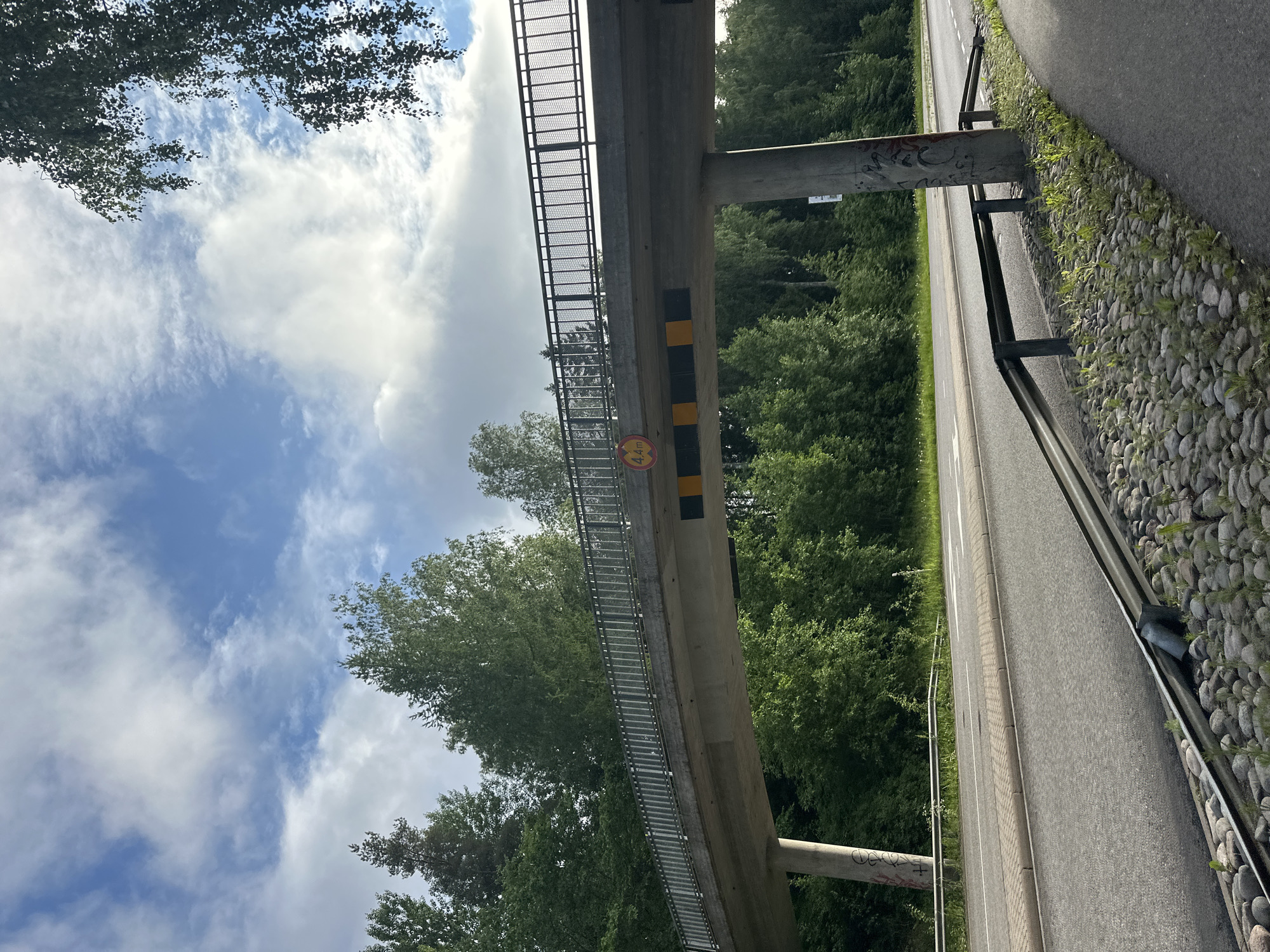 This is a low-angle outdoor photograph of a pedestrian bridge over a road. The bridge is constructed of concrete with a metal railing along its length. Below the bridge, there is a road with two lanes and white lane markings. A guardrail runs along the side of the road. The bridge supports are concrete pillars, one of which has graffiti. The surrounding environment is densely vegetated with trees and green foliage. The sky above is partly cloudy with a mix of blue sky and white clouds. A sign indicating a height restriction of 4.4m is visible under the bridge. The ground in the foreground consists of a paved walkway and an area covered with stones and small plants.