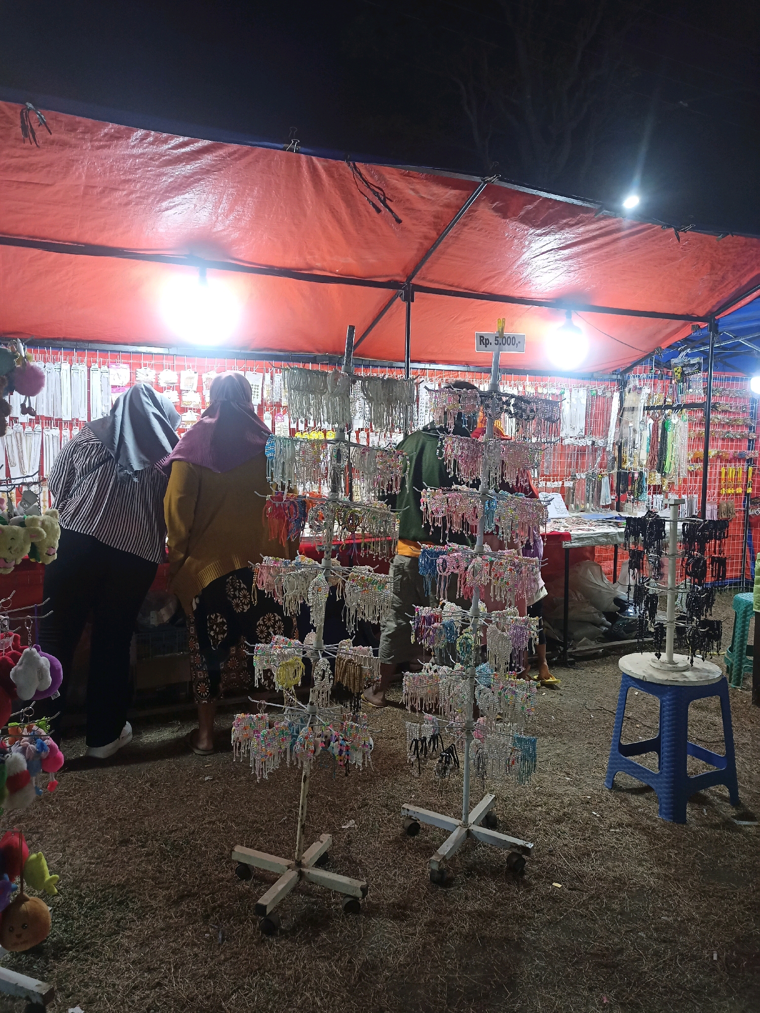 This is a nighttime photograph of a street market stall in Tanon, Indonesia. The stall is covered by a large, reddish-orange tarp supported by metal poles. Bright lights illuminate the scene from beneath the tarp, casting shadows. The stall displays a wide variety of accessories, including keychains, bracelets, and necklaces, hanging from metal racks and grids. A price tag reading "Rp. 5.000,-" is visible. Two women, their faces obscured by hijabs, are browsing the merchandise. One woman is wearing a striped long-sleeved shirt and black pants, while the other is in a mustard-yellow sweater and patterned skirt. In the foreground, a metal display stand with wheels holds colorful bracelets. To the right, a blue plastic stool sits on the ground, which is covered in dry grass and debris. The overall atmosphere suggests a bustling local market at night.
