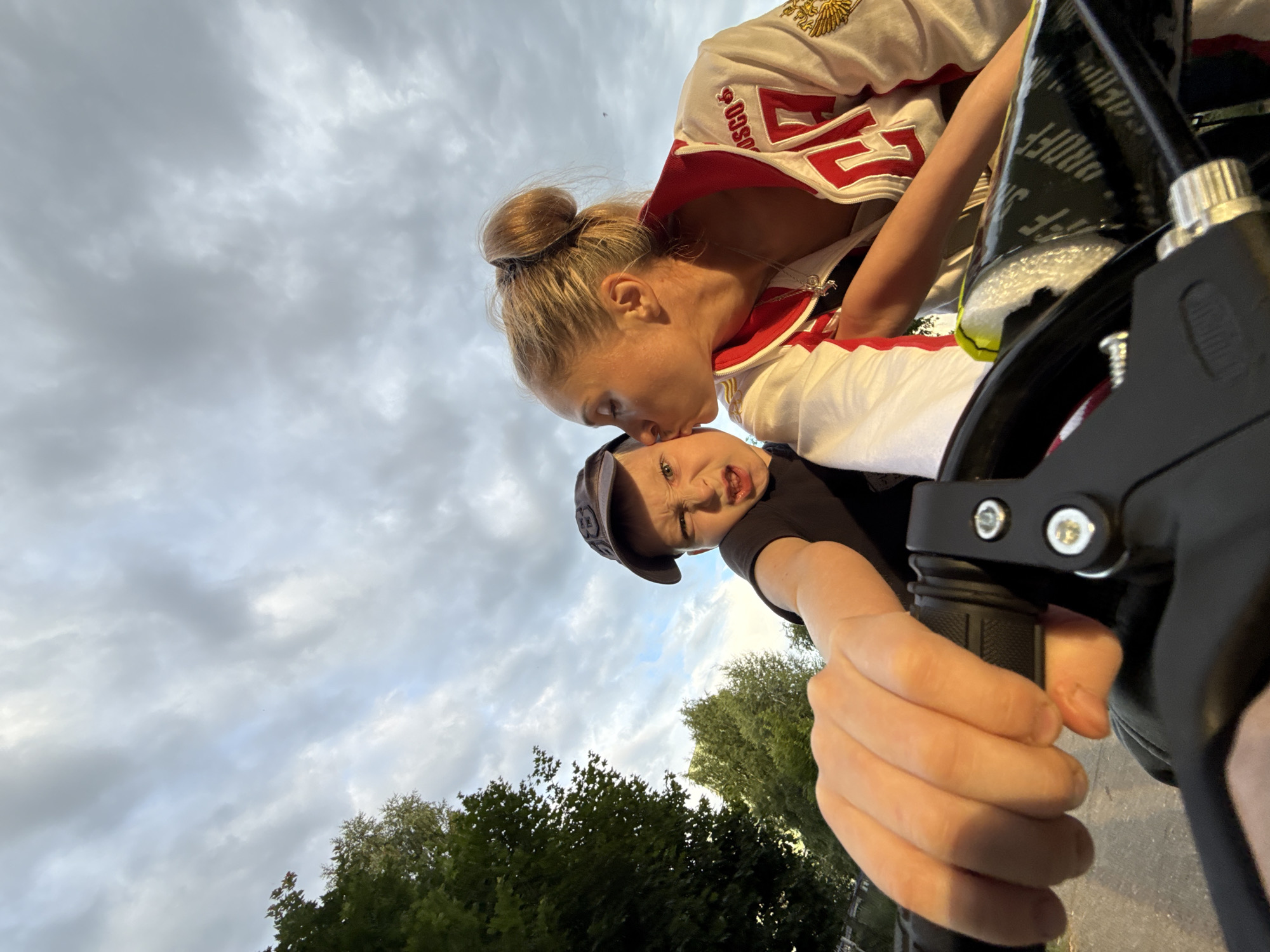 The image shows a young boy wearing a black t-shirt and a baseball cap, with a woman kissing him on the forehead. The woman is wearing a white and red tracksuit with gold embroidery. They appear to be on a bicycle, with the boy holding the handlebars. The background is a cloudy sky with green trees visible at the bottom. The photo is taken from a low angle, looking up at the subjects. The expression on the boy's face suggests playfulness or surprise. The overall mood seems affectionate and joyful. The visible text on the woman's tracksuit is in Russian. The scene is likely outdoors, possibly in a park or a residential area, given the presence of trees. The time of day appears to be late afternoon or early evening due to the cloudy sky and the quality of light.