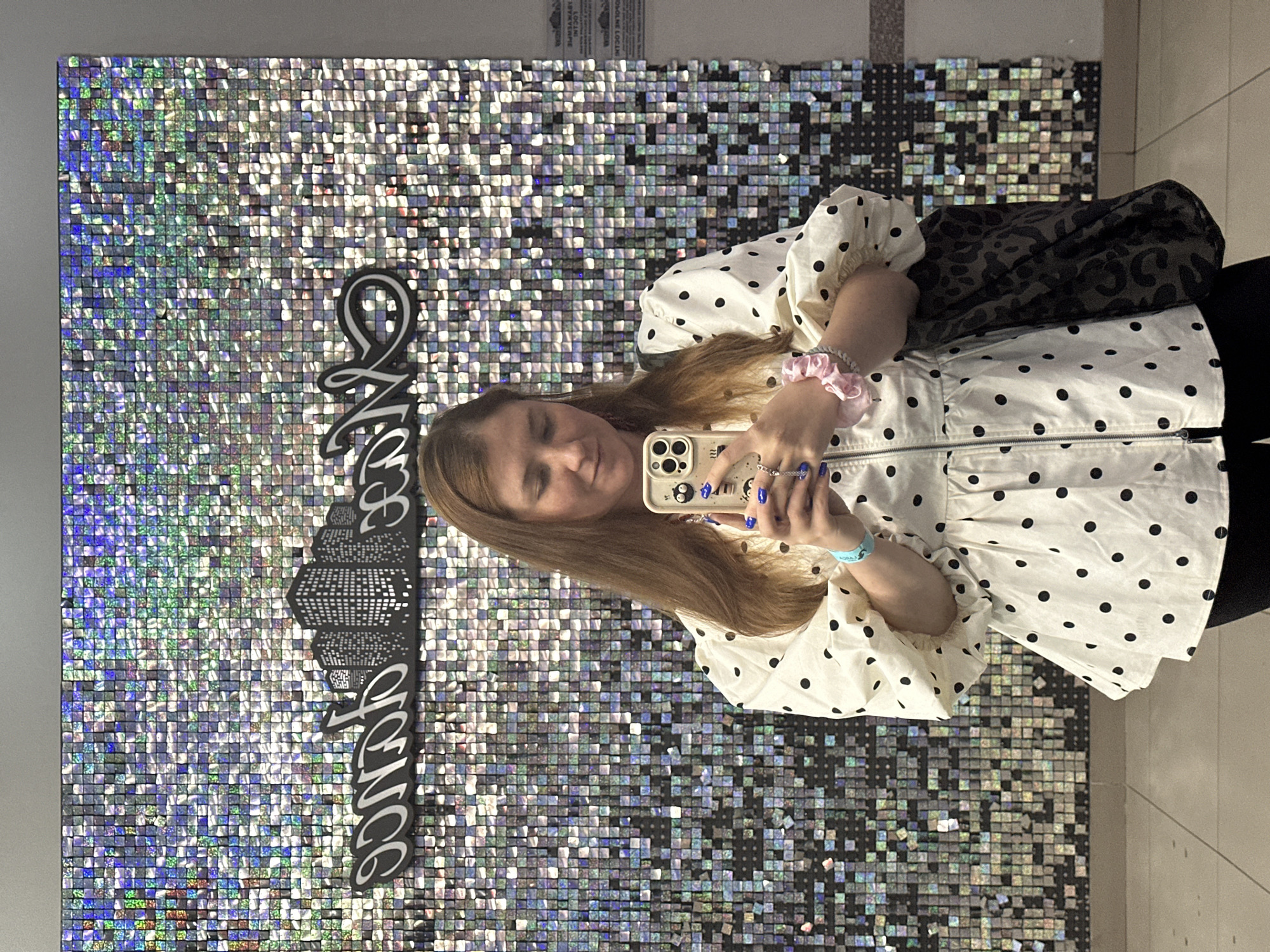 A young woman with long, light brown hair is taking a selfie in front of a wall covered in iridescent squares. She is wearing a white blouse with black polka dots and puffed sleeves, a silver zipper down the front, and black leggings. On her right wrist, she has a turquoise band, and on her left, a light pink scrunchie. She is holding a smartphone with a black and white patterned case in her right hand, and her fingernails are painted bright blue. A dark tote bag with a leopard print pattern hangs from her left shoulder. The wall behind her features a black silhouette of city skyscrapers and cursive lettering. The floor is tiled. The scene appears to be indoors, possibly in a commercial establishment or event space in Petropavlovsk-Kamchatsky, Russia.