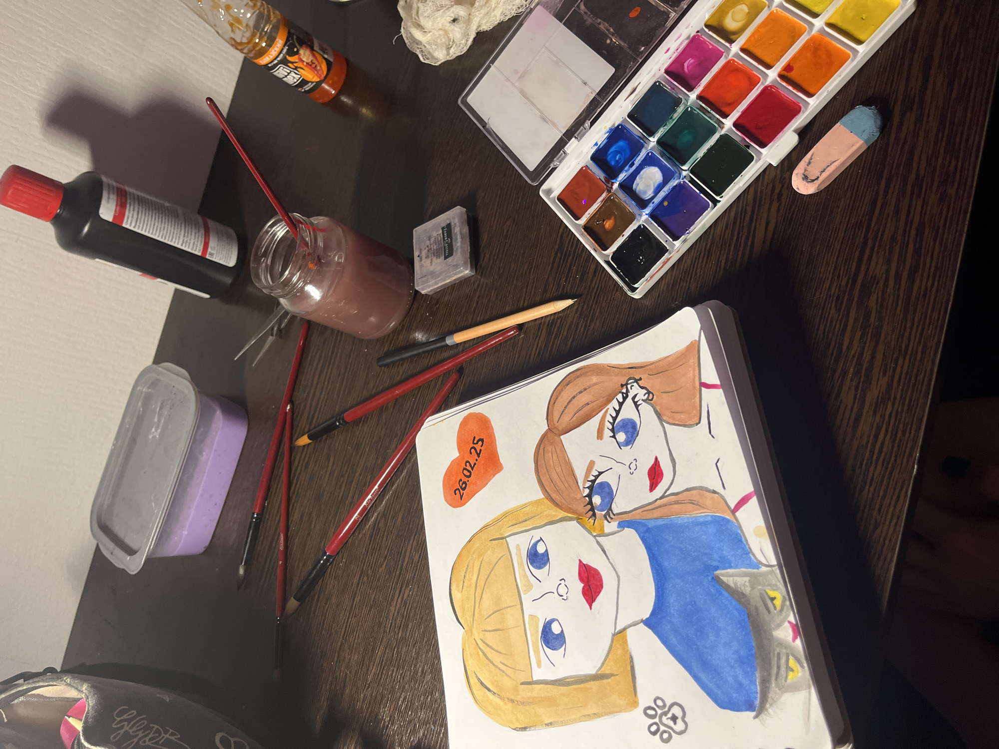 The scene is a desk or table covered in art supplies, seemingly in a residential setting. The predominant color of the table is dark brown. A partially completed drawing of two cartoonish girls is in the foreground. A grey cat head is near the bottom of the drawing. A heart with the date "26.02.25" is drawn on the same paper, at the upper left corner of the drawing. To the upper left of the drawing are several paintbrushes, a grey-lidded container with purple contents, a brown bottle with a red cap, a jar with a reddish-brown liquid and a paintbrush inside, and a bottle of yellow/orange liquid. To the right of these objects sits a small, grey eraser, a plastic watercolor palette with a variety of colored paints. A small white cloth or cotton ball is between the orange liquid and the watercolor palette. The scene lacks any distinct features that could pinpoint it within Saint Petersburg, Russia. No people are visible in the image, the primary subjects of interest are the art materials and the drawing.