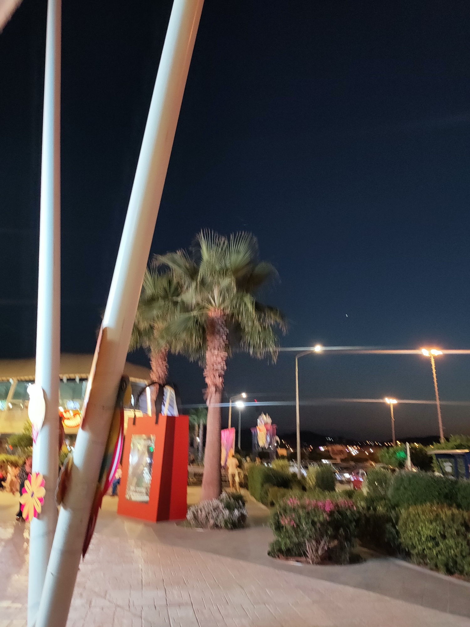 The image shows a nighttime scene outdoors, possibly in a public area or entertainment venue, as suggested by the presence of decorative elements and lighting. Two large, white cylindrical poles dominate the foreground, with a decorative floral element attached to the left pole. Behind the poles, two palm trees are visible, their fronds illuminated from below.
In the middle ground, a bright red, shopping bag-shaped display structure stands prominently. This structure appears to have a transparent section, possibly displaying items within. Further back, more palm trees and lush green bushes and plants are visible, suggesting a landscaped area. Several streetlights with warm yellow illumination are scattered throughout the background, indicating urban or developed surroundings. The sky is a deep, dark blue, consistent with nighttime.
While the prompt mentions Cebalat, Tunisia, there are no explicit visual cues within the image itself to confirm this specific location. However, the presence of palm trees and the architectural style of the lights are consistent with a Mediterranean or North African environment. There are a few indistinct figures of people in the background, too blurred to provide specific details about their actions or interactions. The overall impression is that of an evening gathering or a well-lit outdoor space.