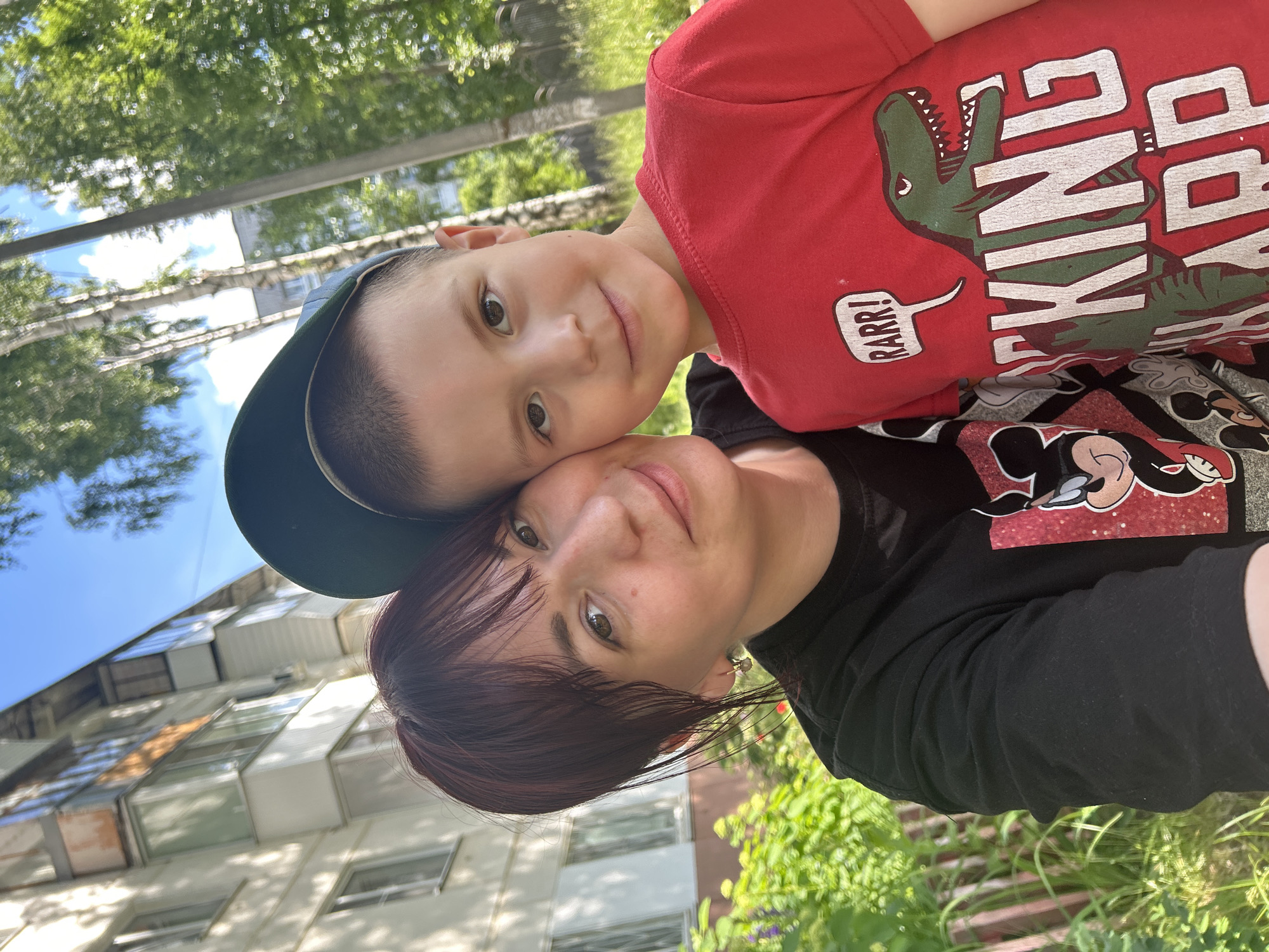 This is a vertically oriented, slightly blurry selfie taken outdoors. The subjects are two people, a child and an adult. The child, positioned at the top of the frame and closer to the camera, has short dark hair and is wearing a blue baseball cap and a red t-shirt. The t-shirt features a graphic of a dinosaur with the text "RARRR!" in a speech bubble and "KING" prominently displayed. The adult, positioned below and slightly further from the camera, has shoulder-length dark hair and is wearing a black t-shirt with Mickey Mouse graphics. Both individuals are looking towards the camera with soft smiles, suggesting a happy and affectionate moment. The background includes green foliage, suggesting a park or garden setting, and a multi-story building with balconies and windows, possibly an apartment building. A bright blue sky is visible through the trees. The lighting suggests it is daytime, likely sunny given the brightness. There are no specific cues to indicate the exact time of day or weather conditions beyond the presence of sunlight. The overall emotion conveyed is one of warmth and closeness between the child and adult. The text visible on the child's shirt reads "RARRR!" and "KING".