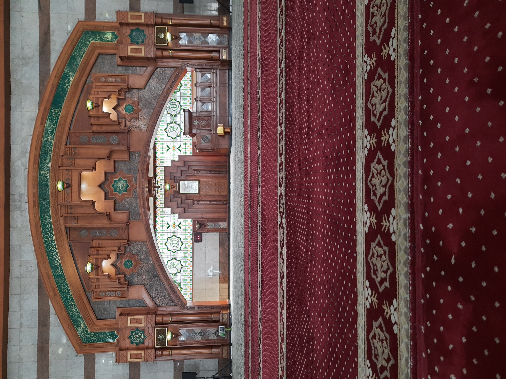 The image shows the interior of a mosque, specifically the prayer hall. In the foreground, a red carpet with a repeating white dotted pattern stretches across the floor, with a decorative border featuring intricate geometric and floral motifs. The carpet is laid out in rows, suggesting the designated prayer spaces.
Behind the carpet, a light-colored carpet or floor covering is visible. Further back, a large, ornate wooden structure dominates the background. This structure features intricate carvings, geometric designs, and is adorned with green calligraphy and what appear to be star-shaped motifs in green. Above this wooden structure, a large arched stained-glass window is visible, filled with green, yellow, and white patterns, including stylized depictions of the Arabic word "Allah" and possibly the name of the Prophet Muhammad.
To the right of the wooden structure, a wooden podium or minbar is present. The walls in the background are a mix of tiled and stone surfaces. The overall impression is one of grandeur and intricate craftsmanship, typical of Islamic architecture.
The image does not contain any people. The scene is static and appears to be a still photograph of the mosque's interior.
While the image is of a mosque interior, the specific location within Bandung City, Indonesia, cannot be determined solely from the visual information provided.