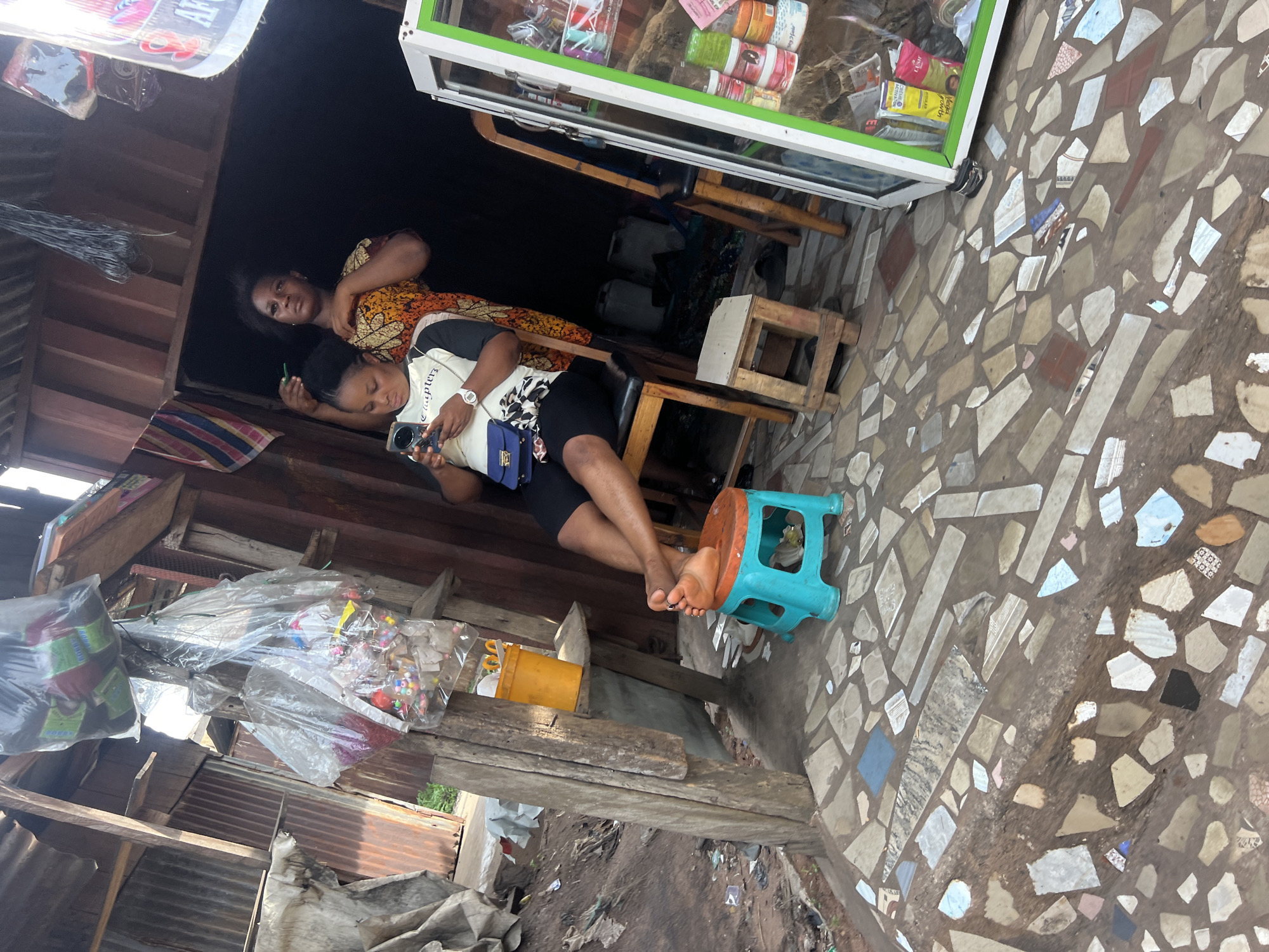 The image depicts two women inside a structure, likely a small shop or stall, in an unknown urban location. One woman, wearing a patterned yellow and orange dress, stands inside the shop looking out. The other woman, in a white shirt with black graphics and black shorts, sits on a wooden chair and stool, holding a mobile phone. The woman sitting has her bare feet propped up on a small blue plastic stool. A display case with merchandise is visible along the wall. The flooring is mosaic tiled, and the surrounding area outside the shop appears to be cluttered with debris and miscellaneous items. The structure itself appears to be made of wood and corrugated metal.